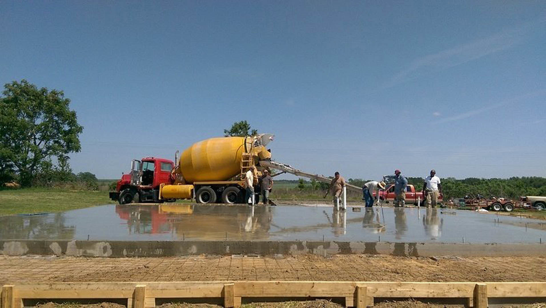 Men standing beside a large yellow concrete mixer truck on a construction site with trees and sky in the background