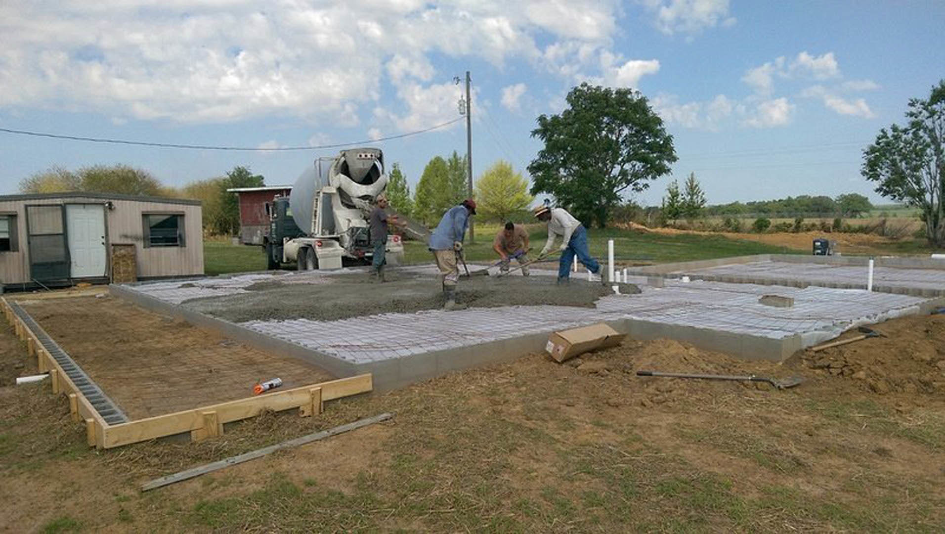 Men pouring and leveling concrete slab foundation outdoors, cement mixer nearby, grassy yard with trees, white doors leaning against wall in background