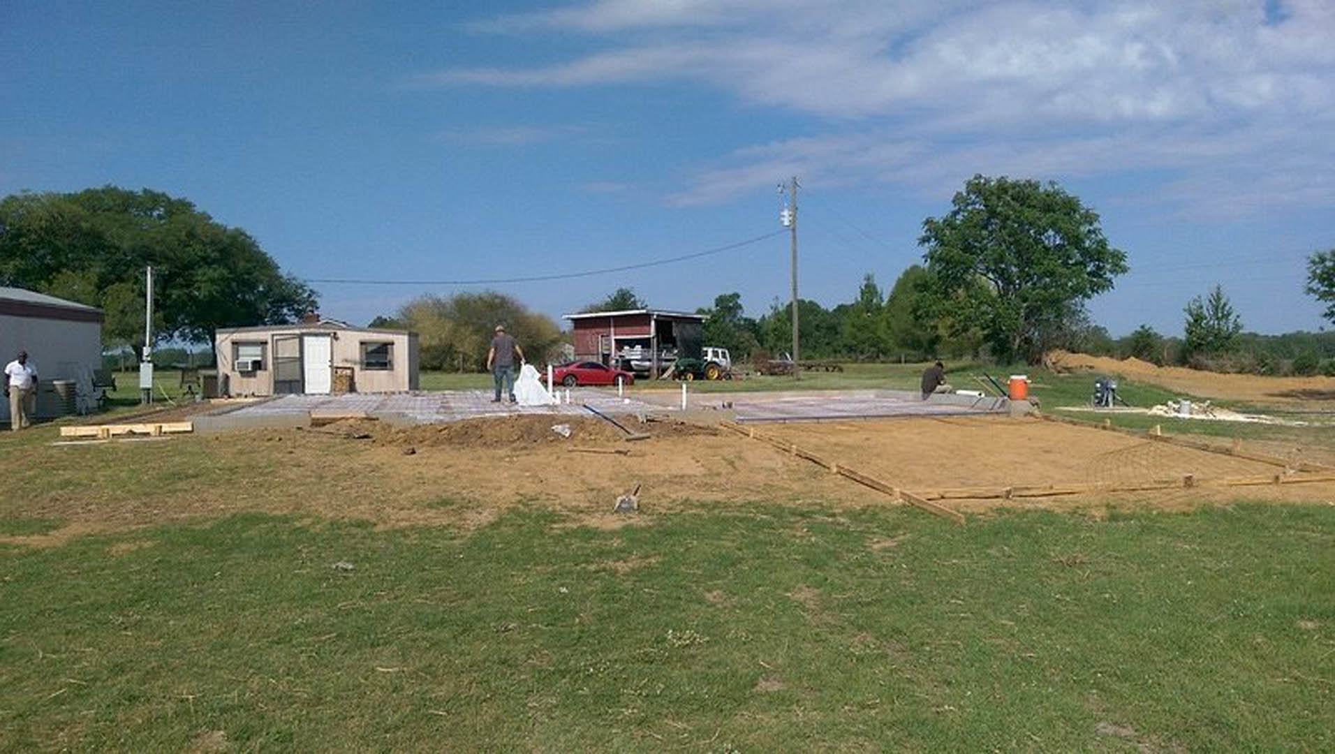 Concrete foundation under construction with two workers, grassy lot, white door on adjacent building, garage door, leafy tree, partly cloudy sky
