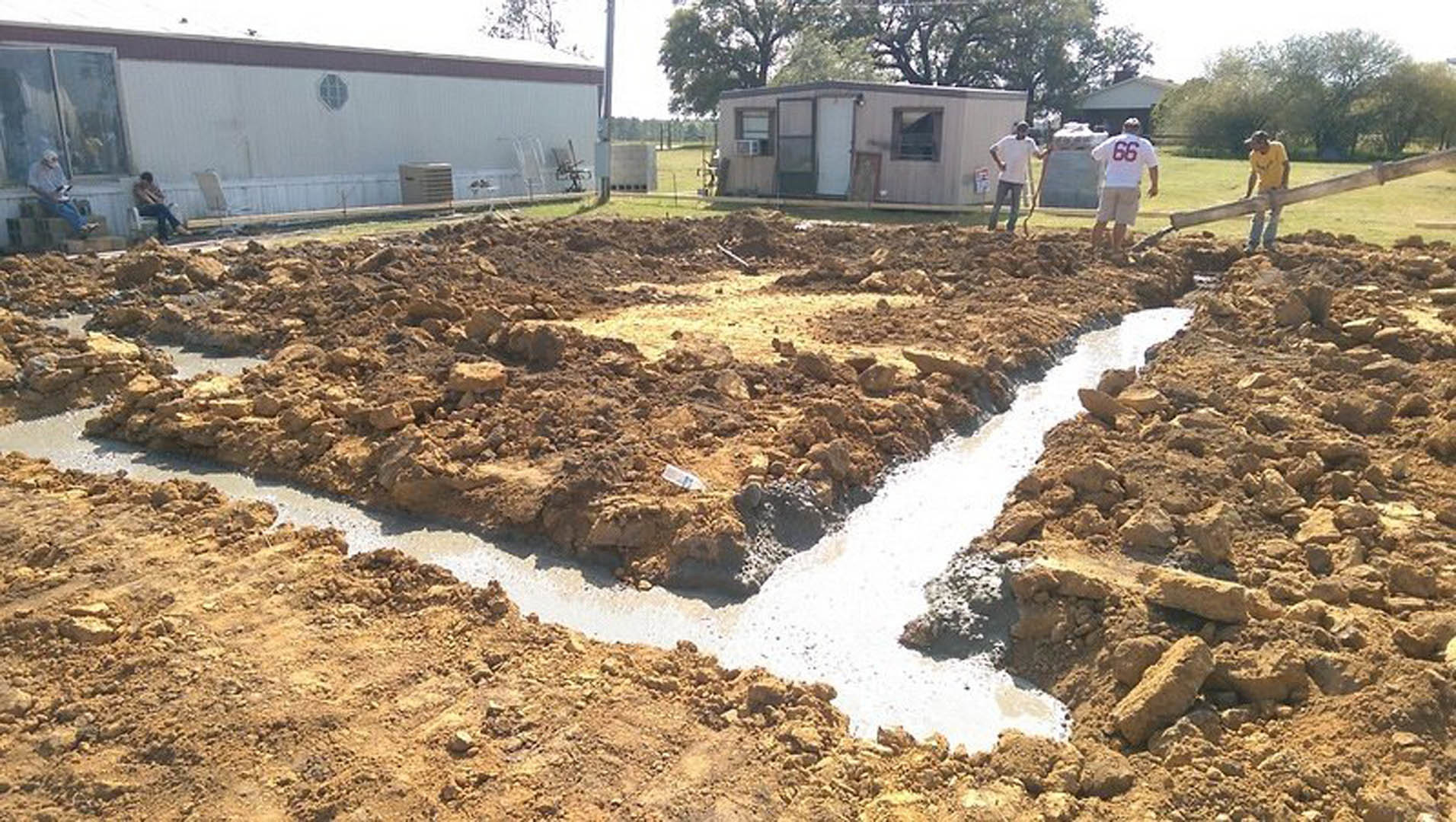 Several men standing on bare dirt in front of a partially visible custom home exterior, surrounded by trees and grass under a clear sky
