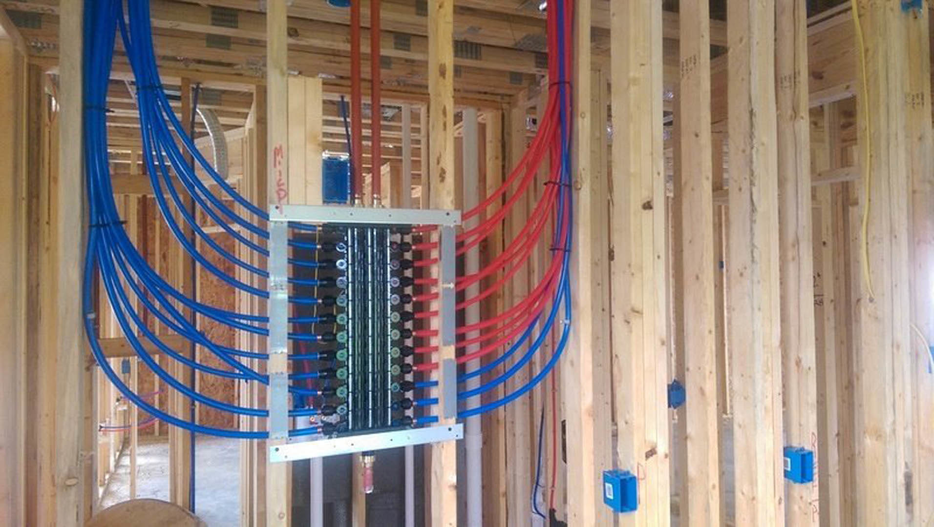 Exposed blue and red plumbing pipes and electrical wires running through unfinished wood framing in a utility room