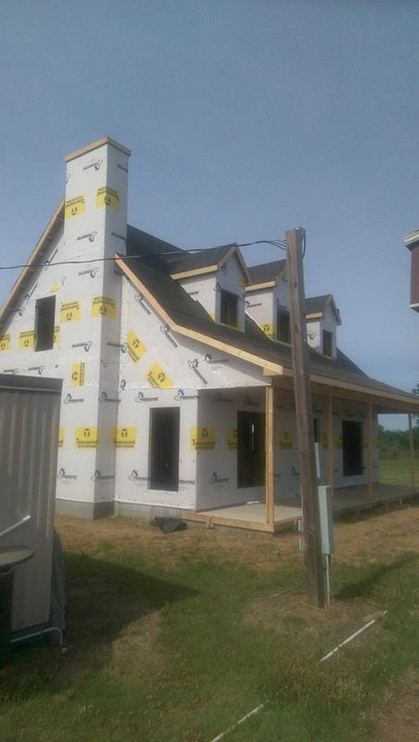 Partially built house with yellow and black insulation panels, exposed chimney, white pipe along exterior wall, grassy yard, and cloudy sky