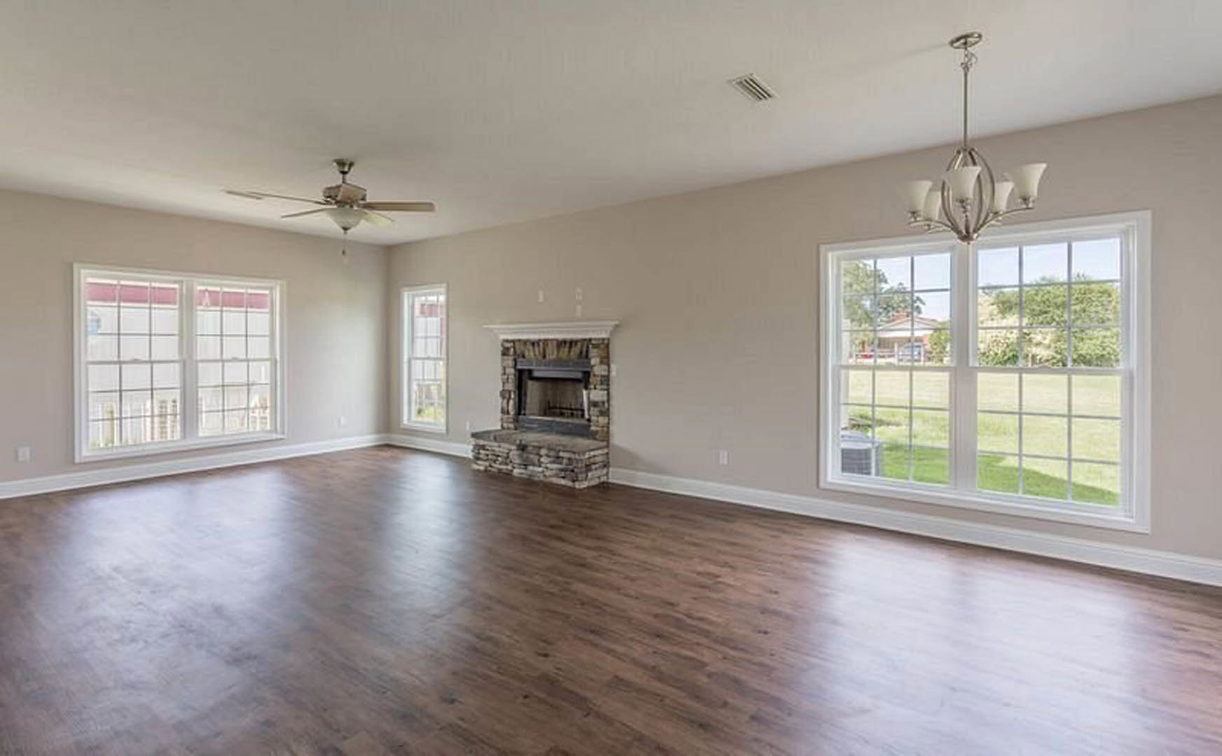 Spacious living room with hardwood floors, stone fireplace, ceiling fan with light, large window overlooking grassy yard and trees, plaster walls