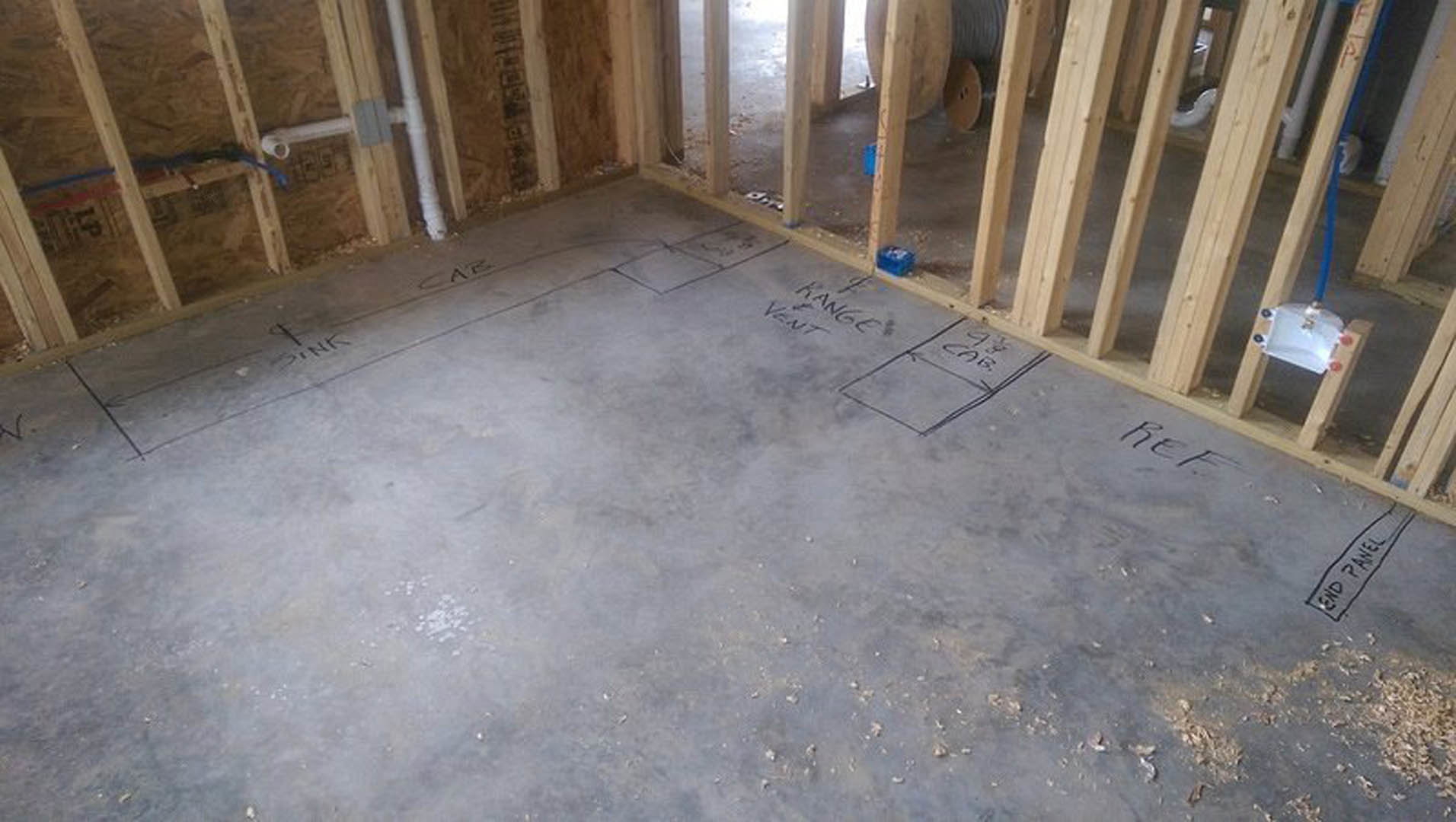 Concrete floor with hand-drawn markings, surrounded by unfinished wood framing and construction materials in a residential interior remodel.