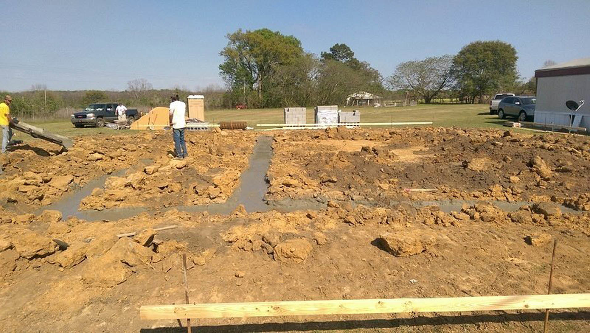 Man standing on muddy construction site near wooden board, with parked truck and car in background, surrounded by bare soil and scattered leaves.