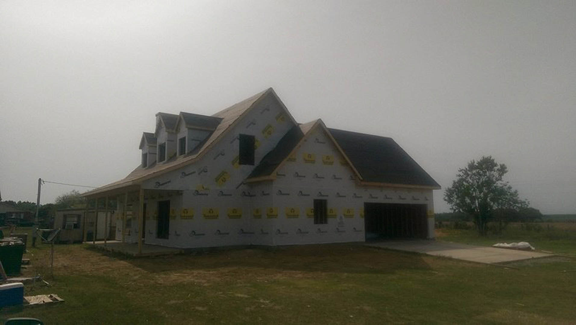 Partially built house with exposed wooden framing, black roof, attached garage, yellow and black siding, construction materials scattered on grassy lot, tree in background