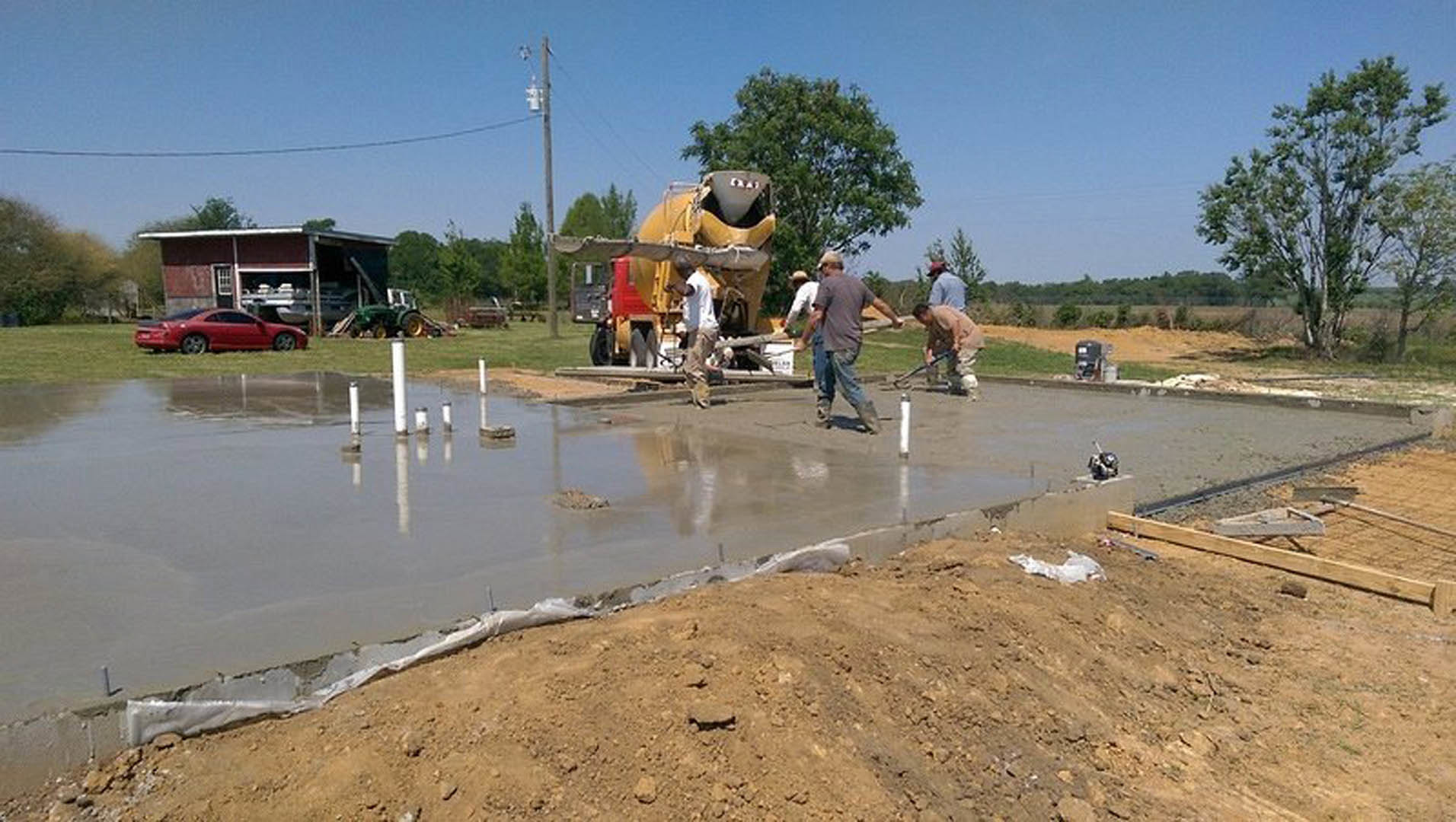 Workers pouring and leveling concrete slab foundation beside parked red car, cement mixer, and muddy ground with scattered construction tools and trees in background