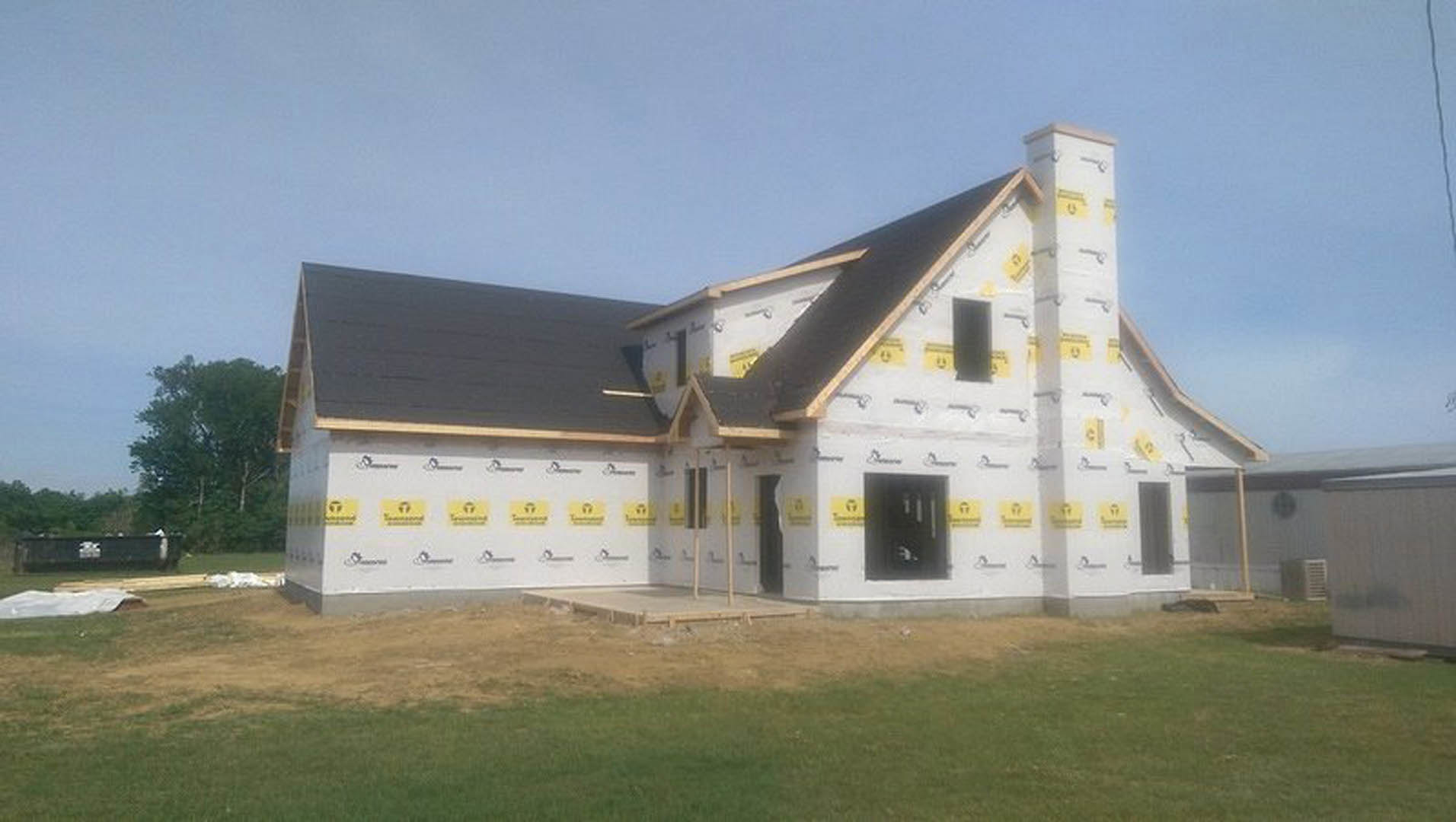 White house under construction with yellow tape along exterior wall, grassy yard, wooden fence, and trees in background