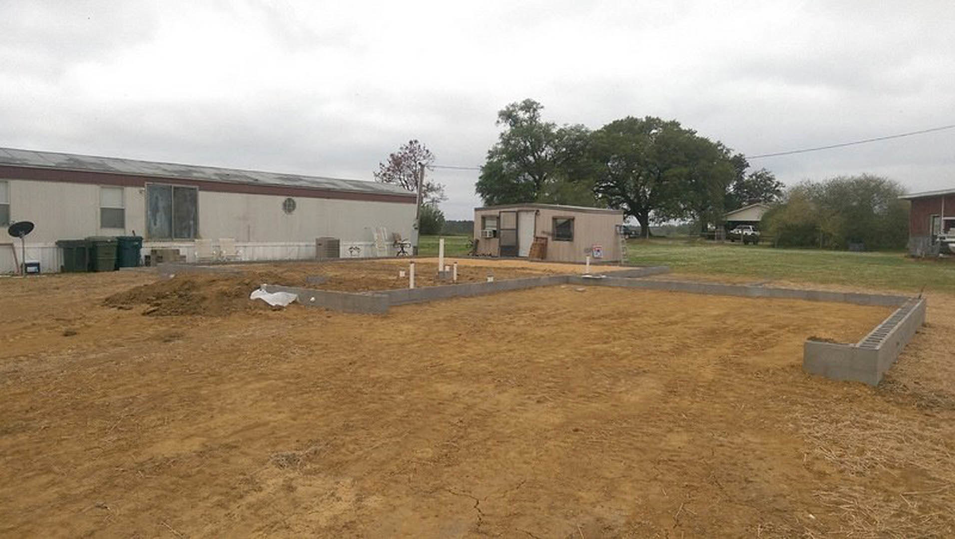 Partially constructed home with exposed concrete foundation, white drainage grate, dirt lot with white poles, small white shed with window, man standing near building, surrounding