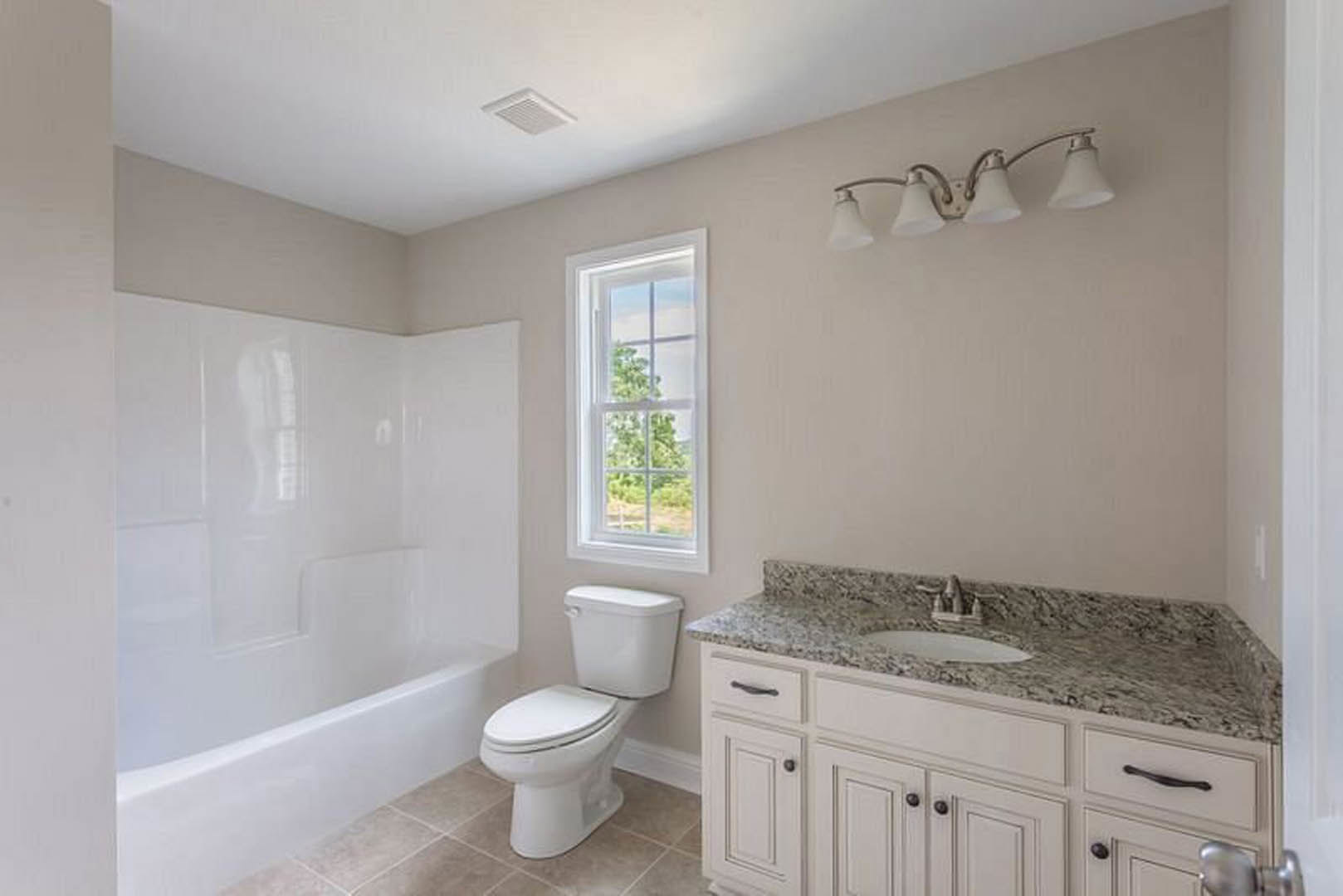Bathroom with white toilet and marble countertop sink, two-lamp light fixture above, window showing tree outside, tiled walls and cabinetry.