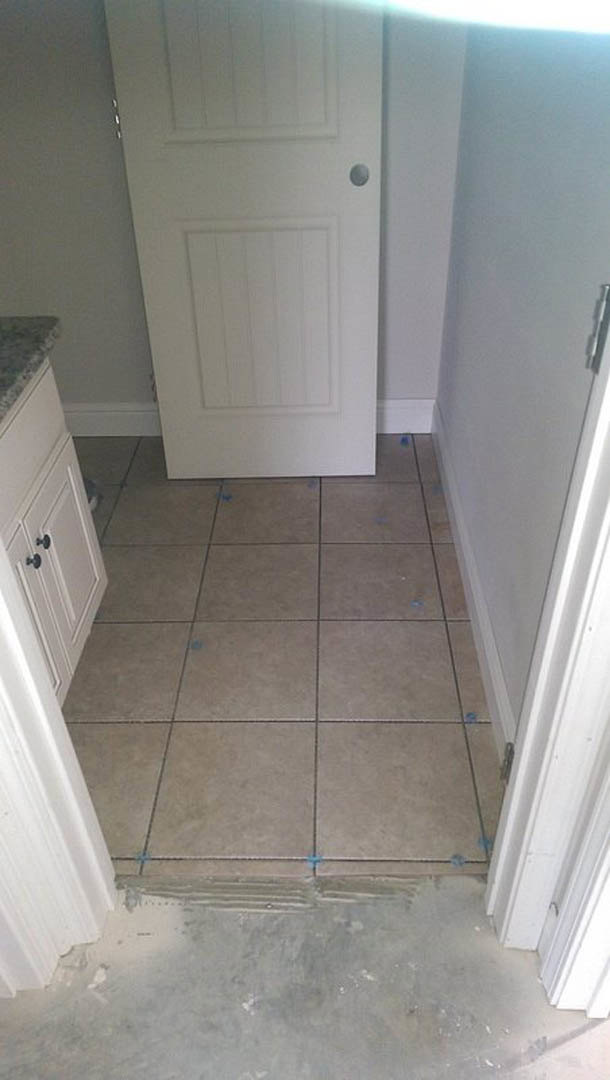 White door with round knob in a kitchen, tile flooring with blue tape, plaster walls, and concrete floor visible in foreground.