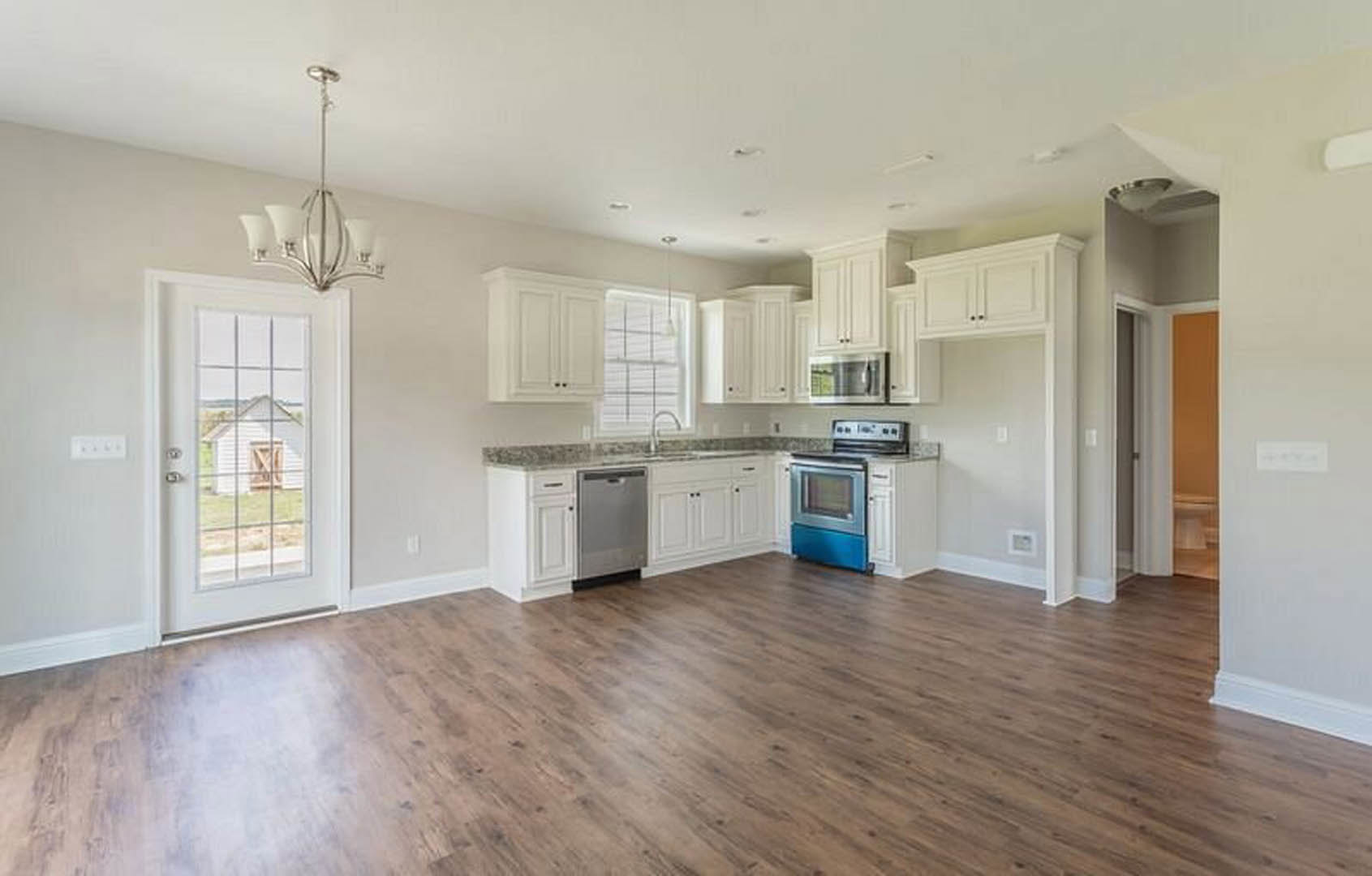 Open kitchen and dining area with wood flooring, white cabinetry, stainless steel stove and microwave, modern chandelier, and a white door offering a view of a barn outside.