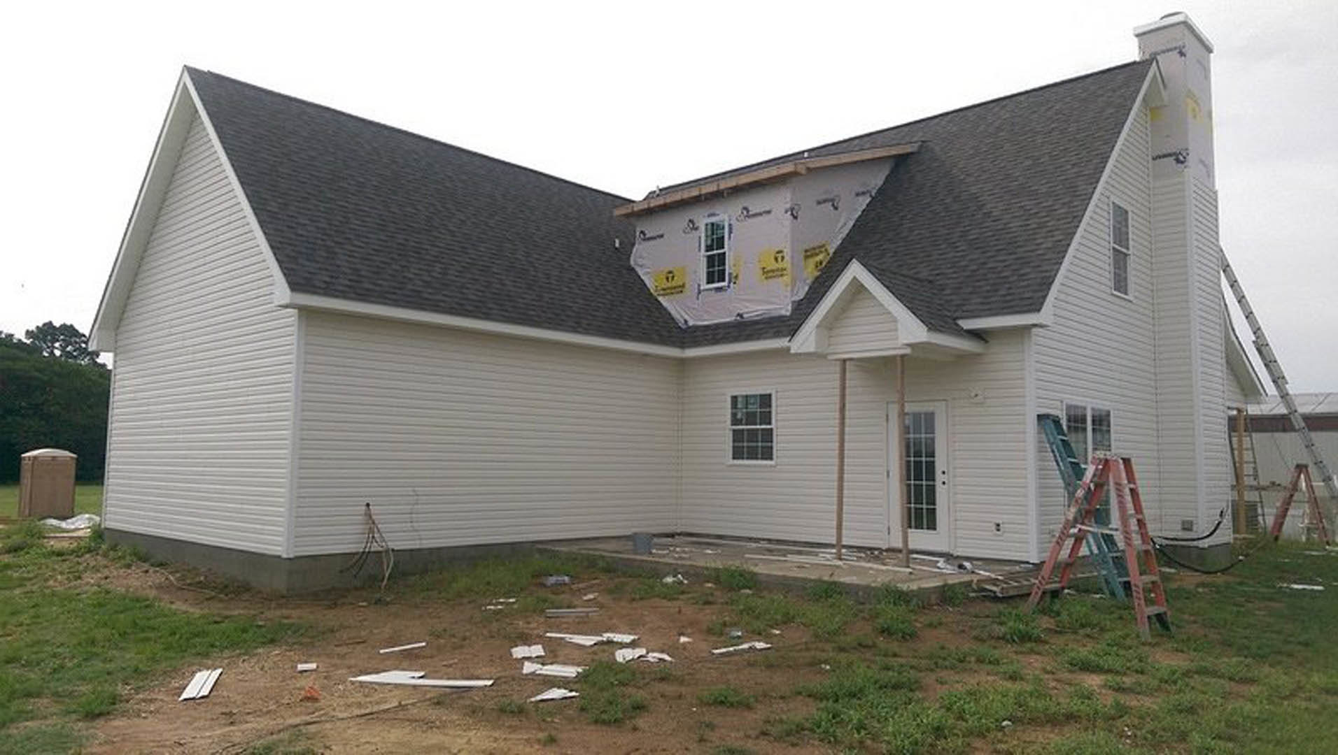 Partially constructed house with finished shingle roof, multi-pane window, white siding, and ladders leaning against exterior walls, surrounded by grass.