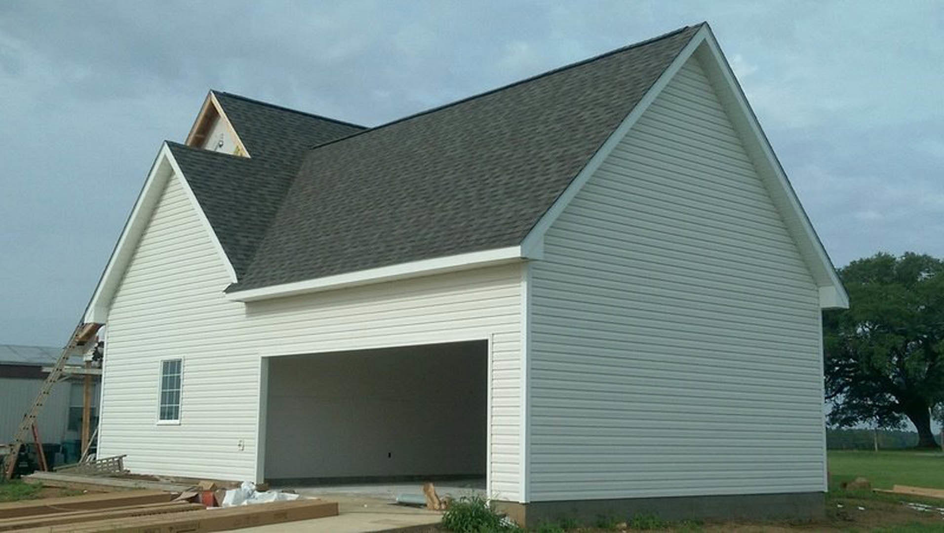 White garage with paneled door, gray shingle roof, white siding, tree with green leaves beside driveway, ladder leaning against exterior wall, man standing near garage entrance