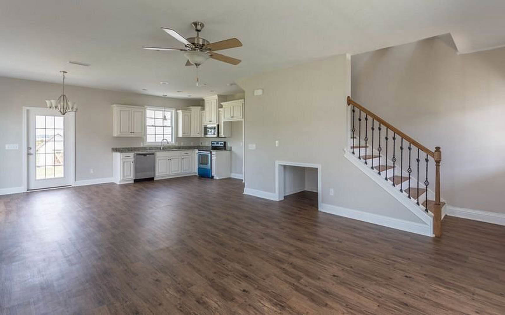 Open living area featuring wood flooring, white walls, wooden staircase with matching railings, ceiling fan with light fixture, and a white door with glass window.