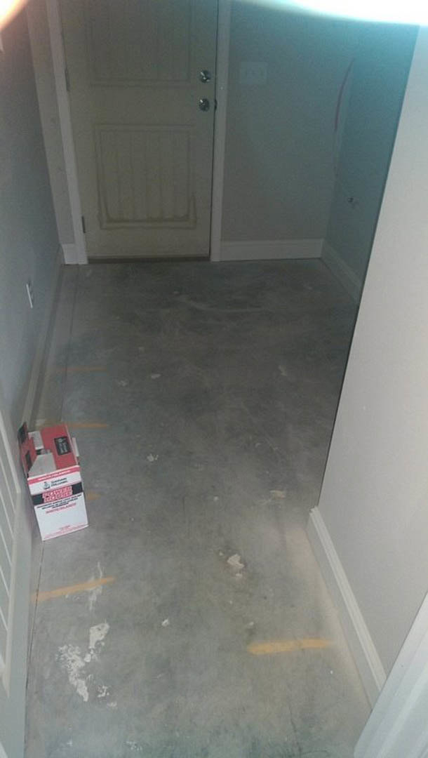 Concrete floor with a cardboard box, white paneled ceiling, white door with silver knobs, close-up of a grey surface, plaster walls, refrigerator in the background.