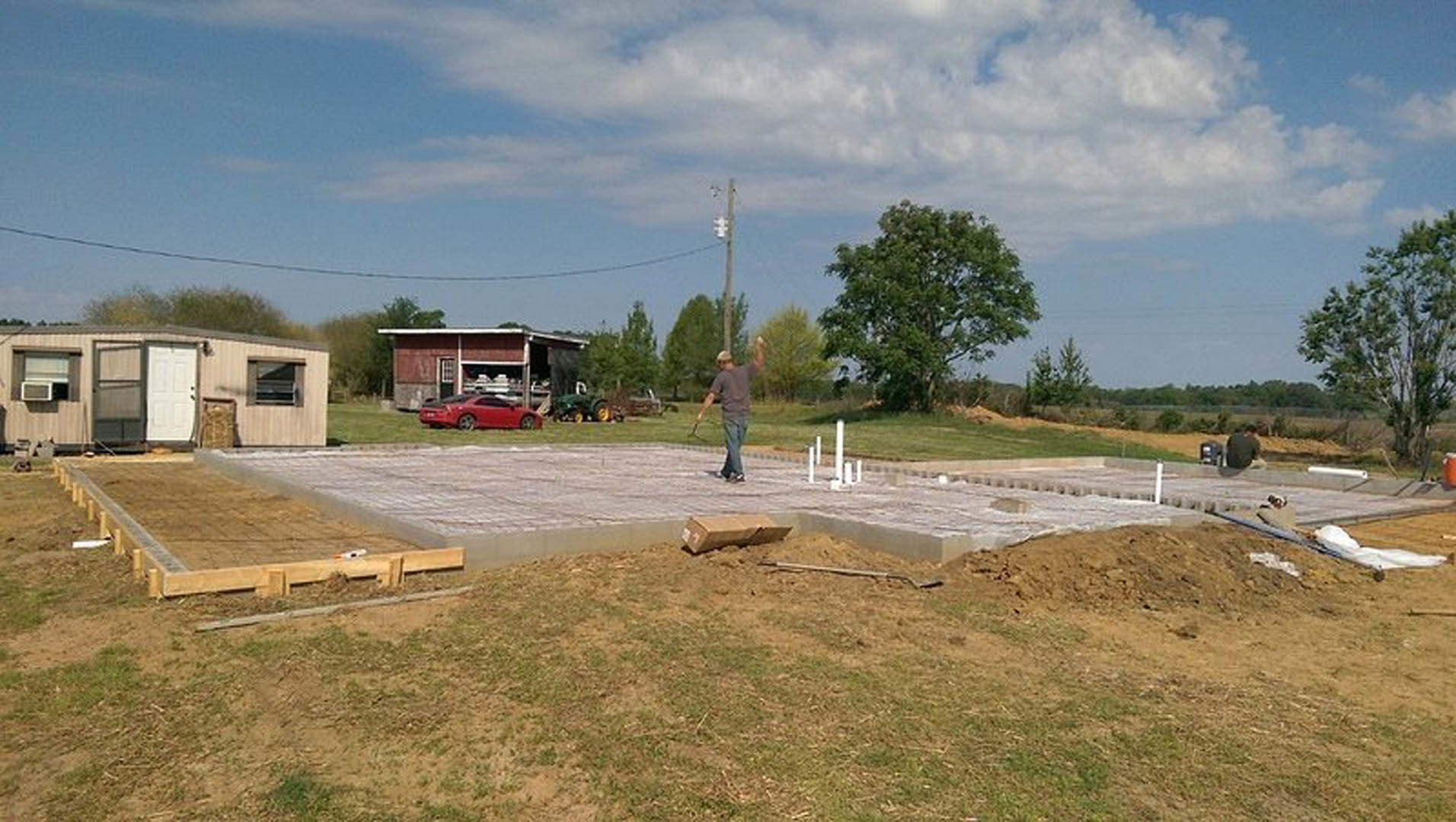 Man standing on unfinished concrete slab in front of a white garage door, leafy tree and shed visible, grassy lot and cloudy sky in background