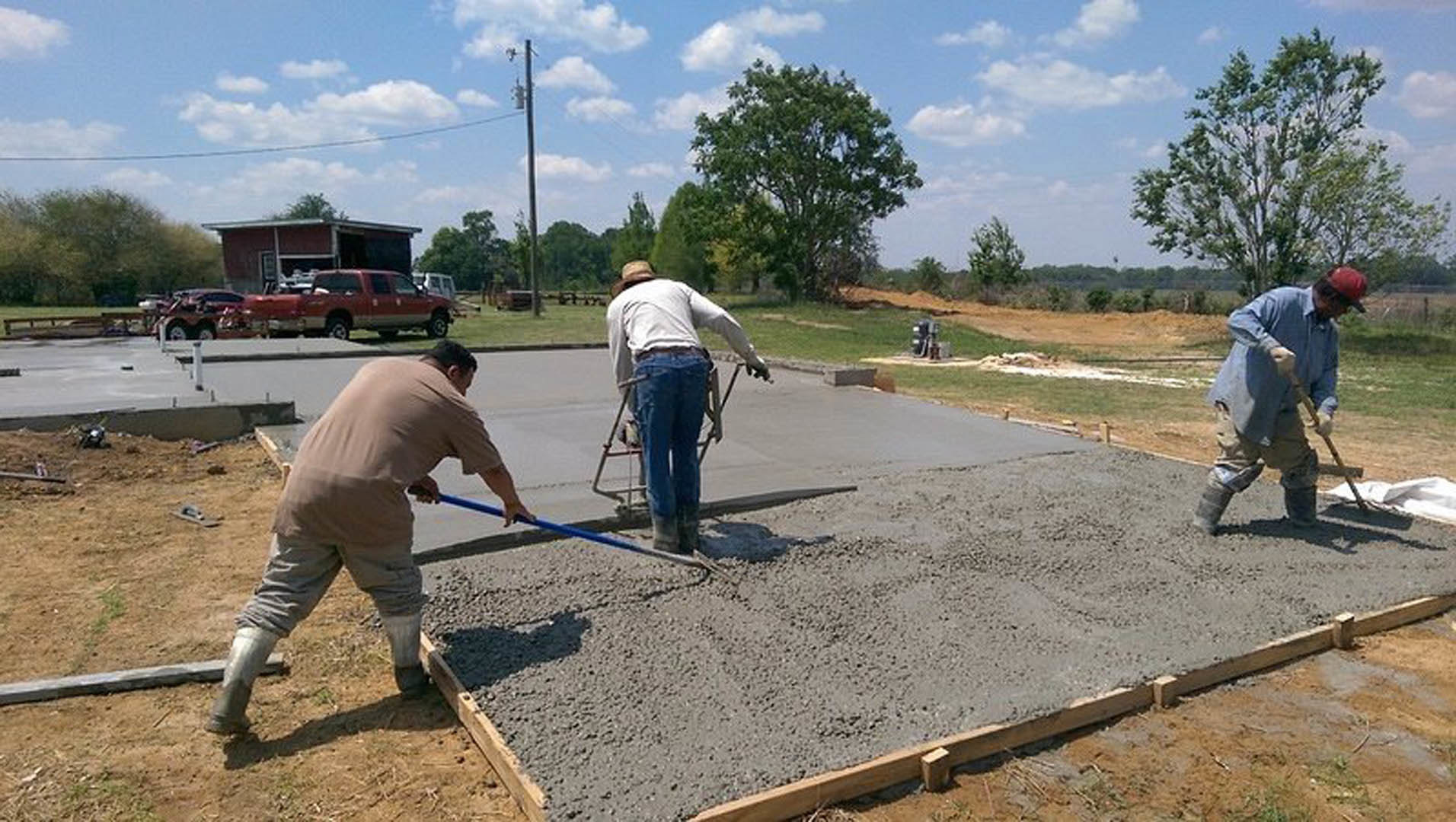 Two men leveling freshly poured concrete in a driveway, one using a shovel and another with a walker nearby; a red truck parked in the background, trees and blue sky visible.