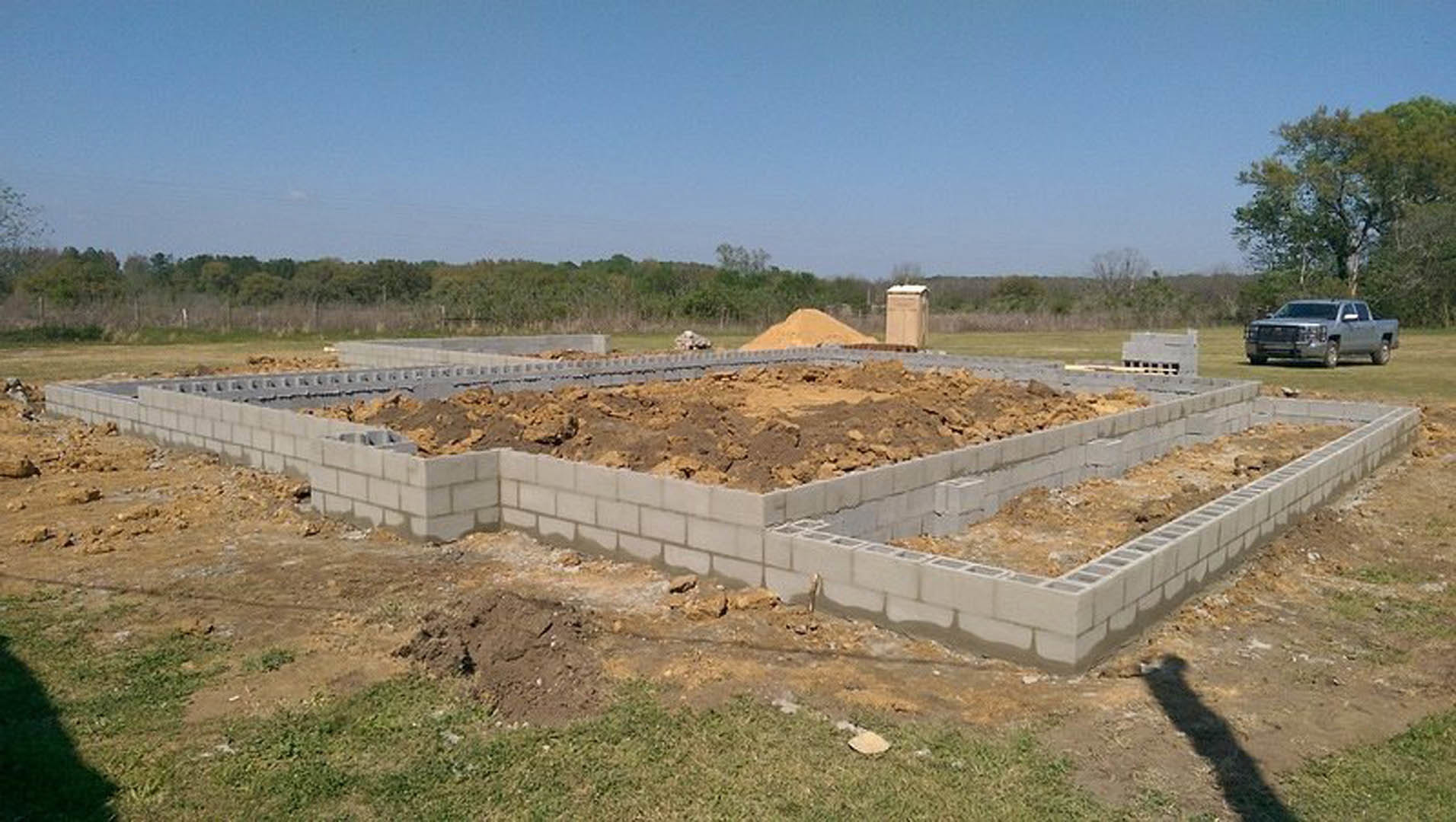 Concrete house foundation surrounded by soil and grass, silver pickup truck parked nearby, trees and blue sky in background, shadow of person cast on ground