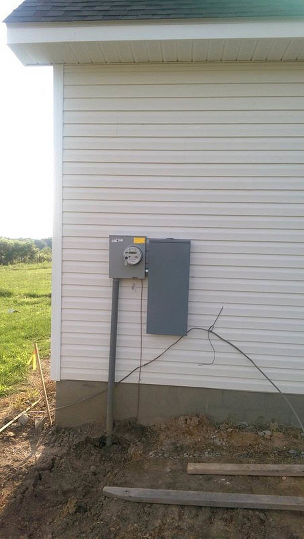 Grey electrical box mounted on light-colored siding, wooden planks and green grass at base of exterior wall, window partially visible.