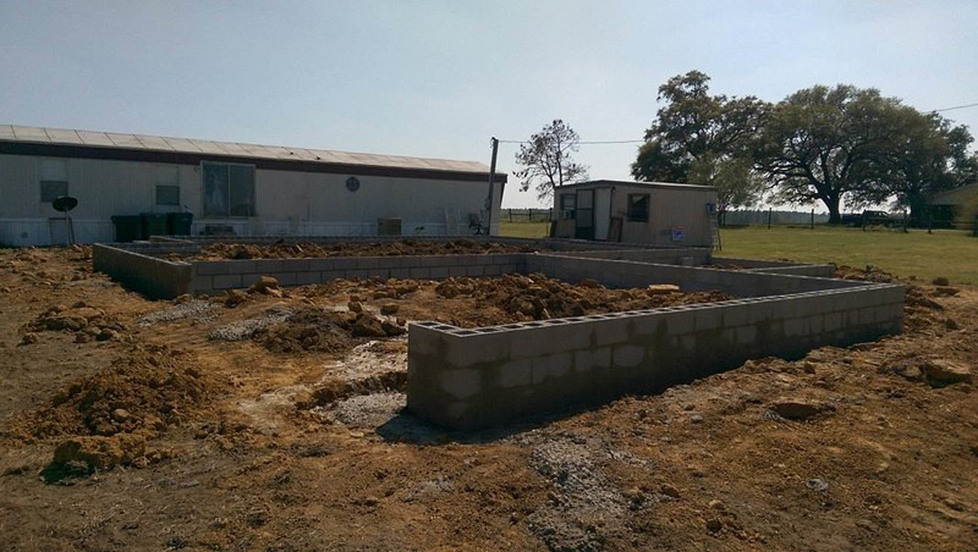 Dirt-covered building site with exposed foundation, surrounding trees, and partially constructed house featuring square-framed window