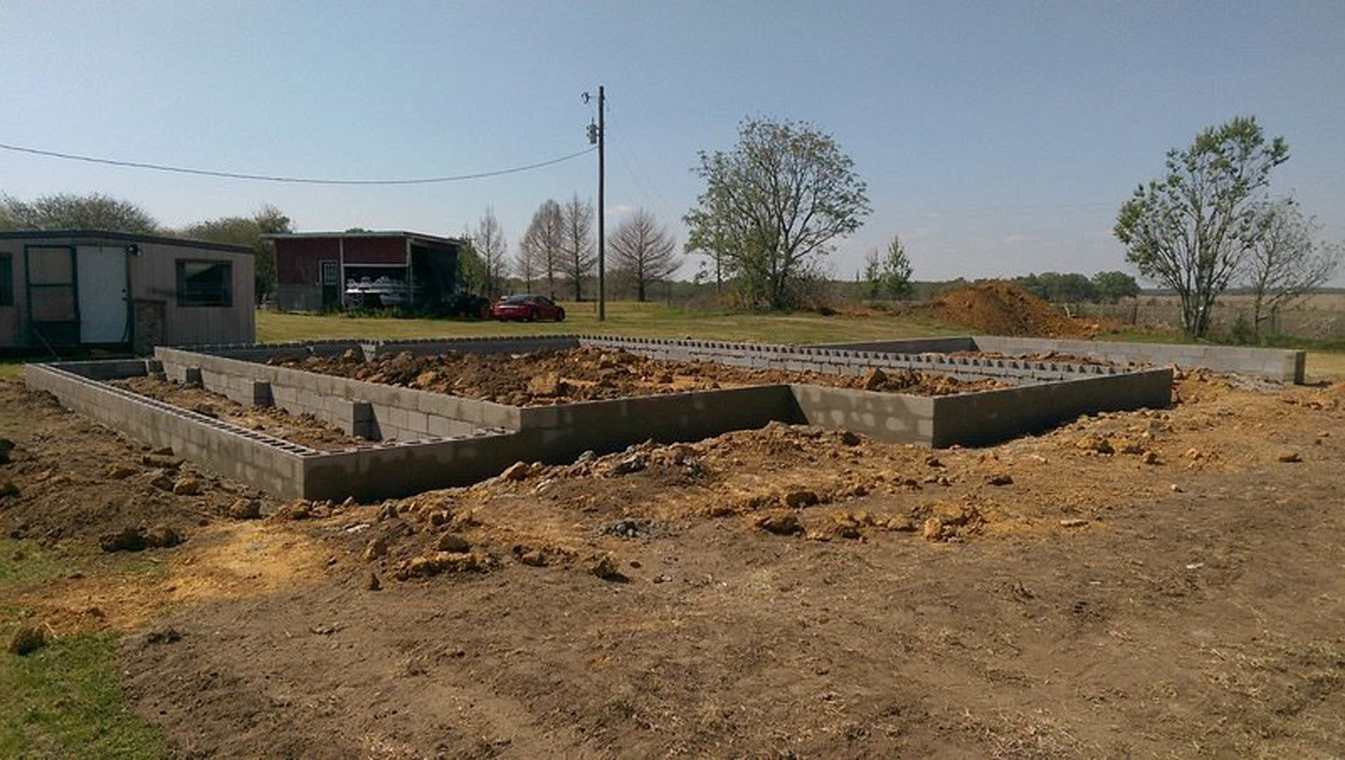 Dirt-filled construction site with exposed soil foundation, leafless tree, grassy field, and partially built house featuring a garage door and trailer