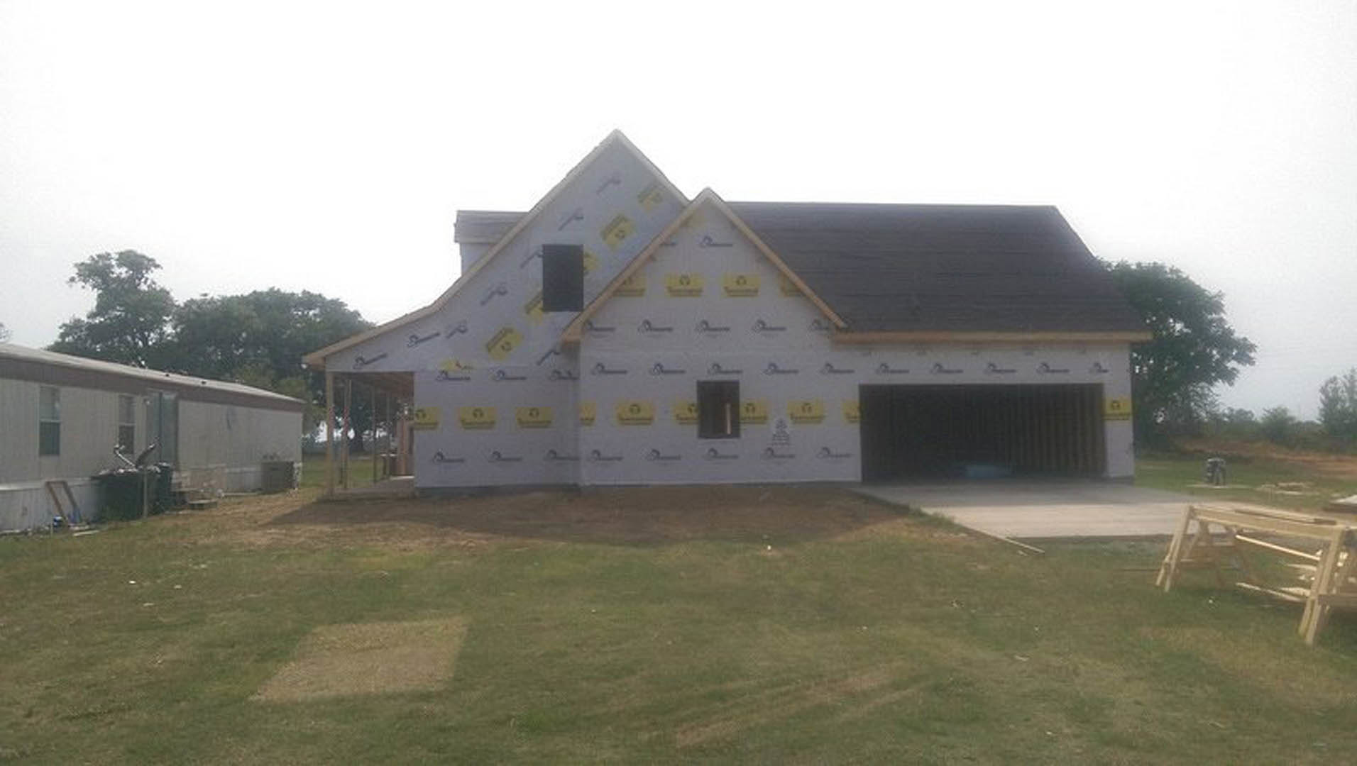 Two-story house with gray siding, attached two-car garage, shingle roof, white-framed windows, and manicured green lawn bordered by young trees.