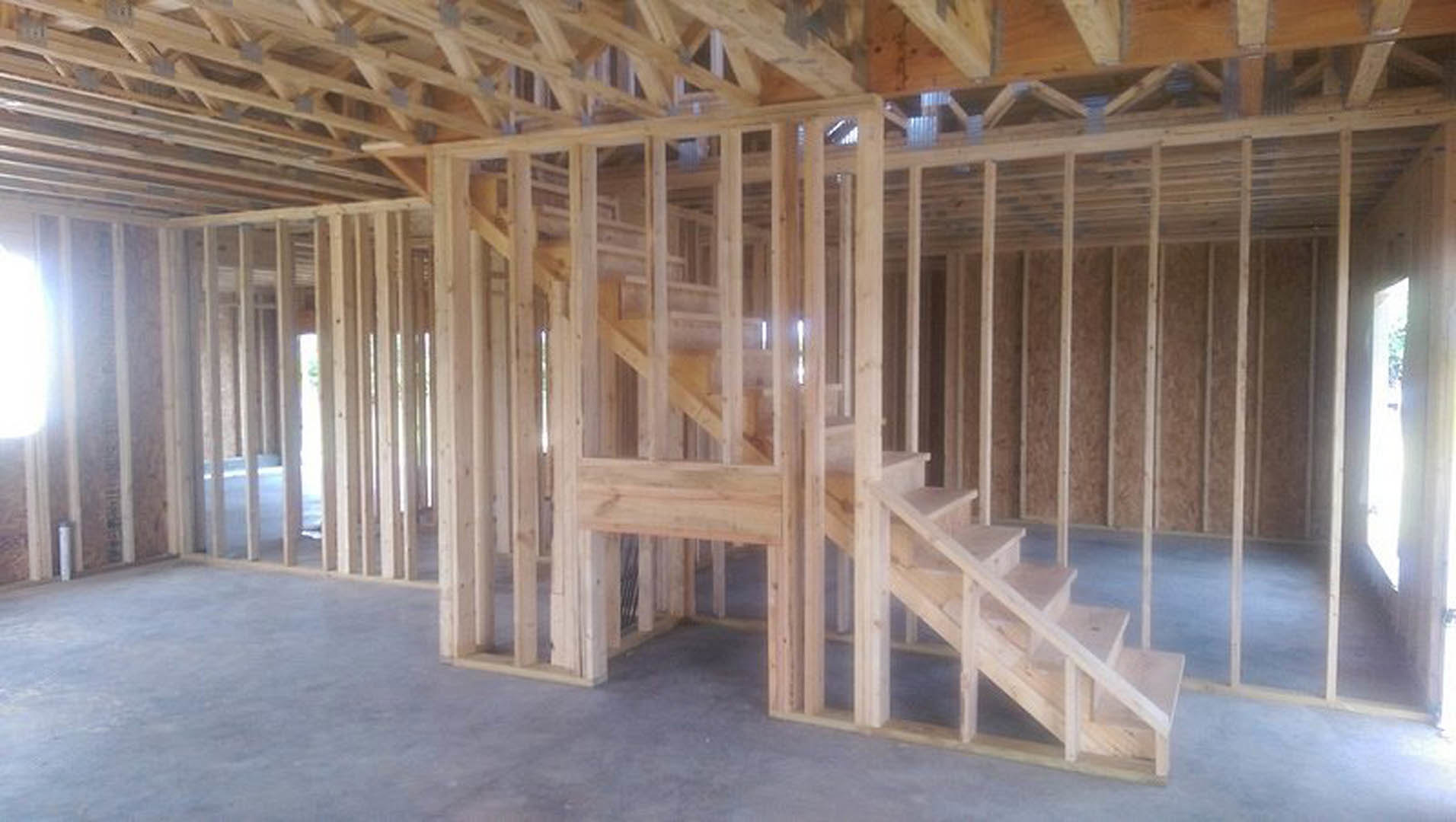 Wooden staircase with natural hardwood treads and risers, surrounded by exposed wood framing and beams, set against white walls with ceiling insulation visible.