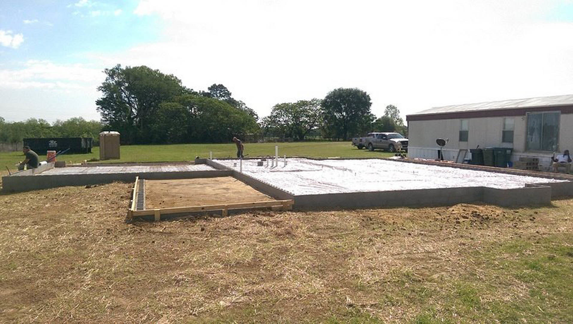 Modern two-story home with light-colored siding, large windows, and a landscaped front yard featuring green grass and mature trees; man standing near entrance, trash cans visible