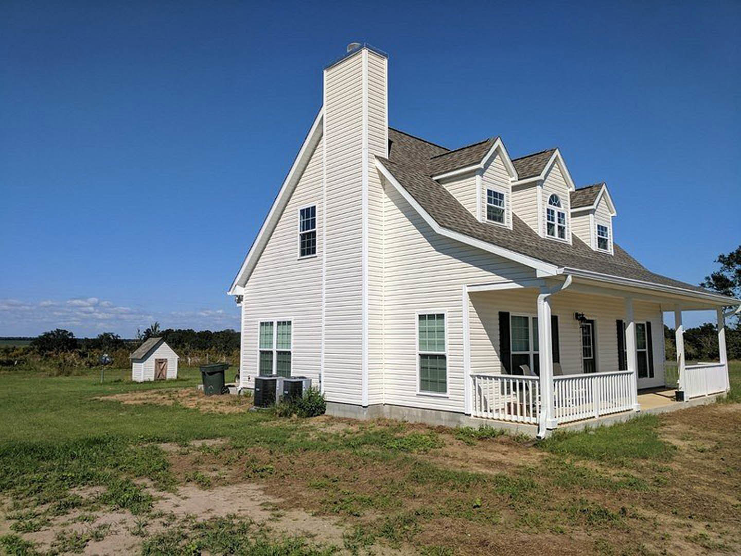 White farmhouse with multi-pane windows, covered porch featuring white railings, manicured lawn, and small white shed in grassy field under blue sky