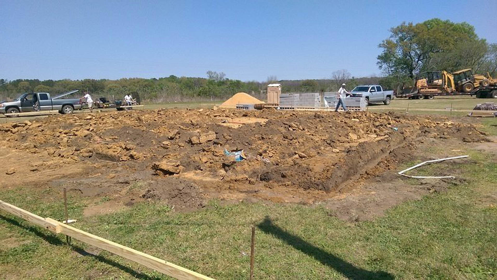 Large pile of soil on grassy lot with white truck and tractor parked nearby, trees and sky in background, person walking in foreground.