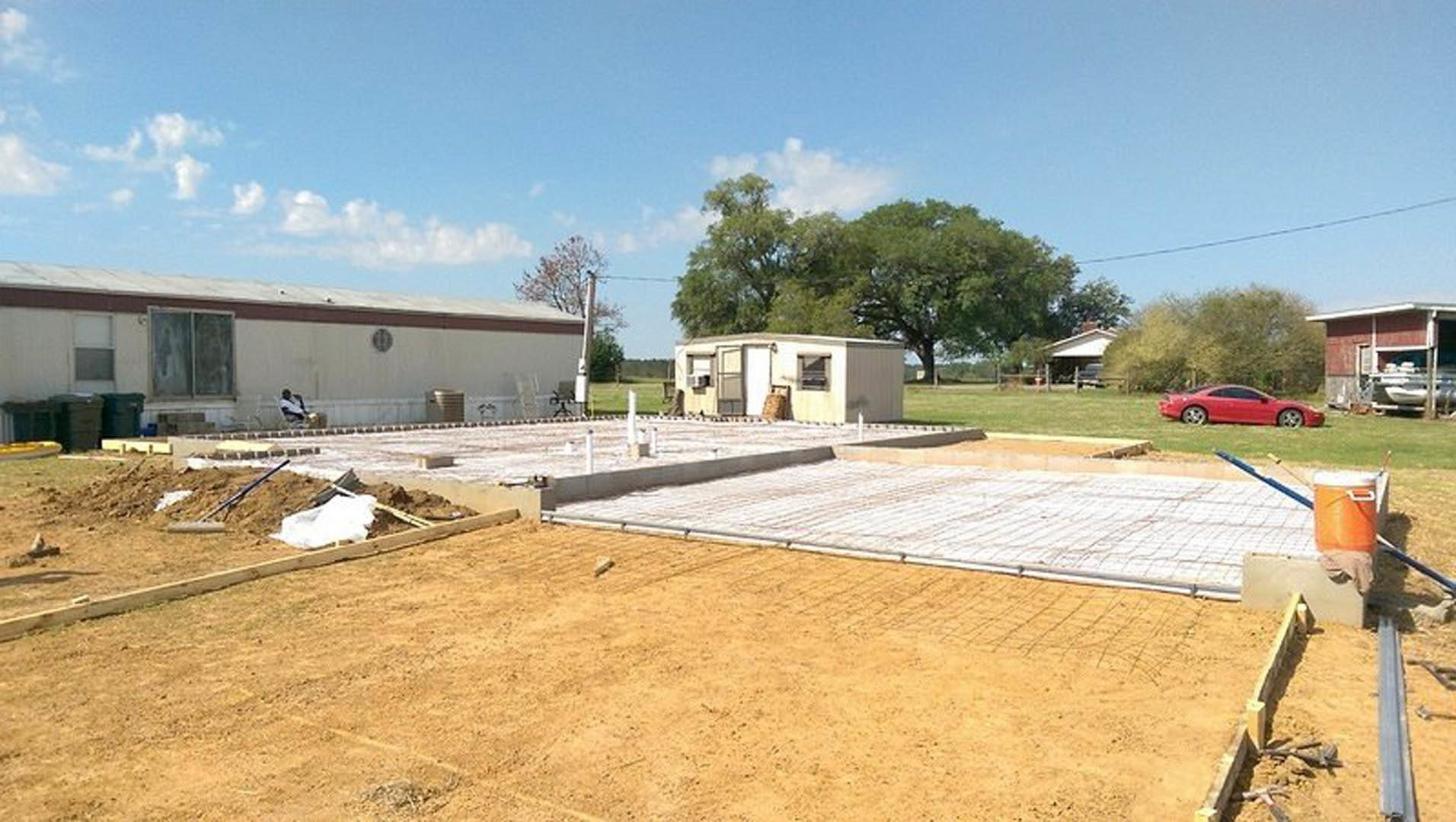 Partially built house with exposed framing and foundation, adjacent to a metal shed and white trailer, grassy lot with scattered construction materials, red car parked nearby, man
