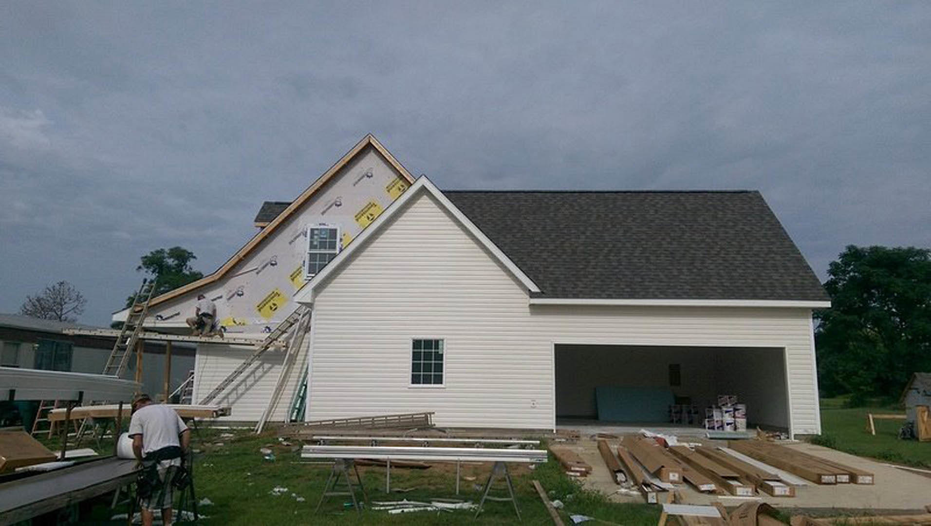 Partially built house with exposed framing, attached garage, metal railings on grassy yard, tree with green leaves, close-up window, and several cardboard boxes scattered outside