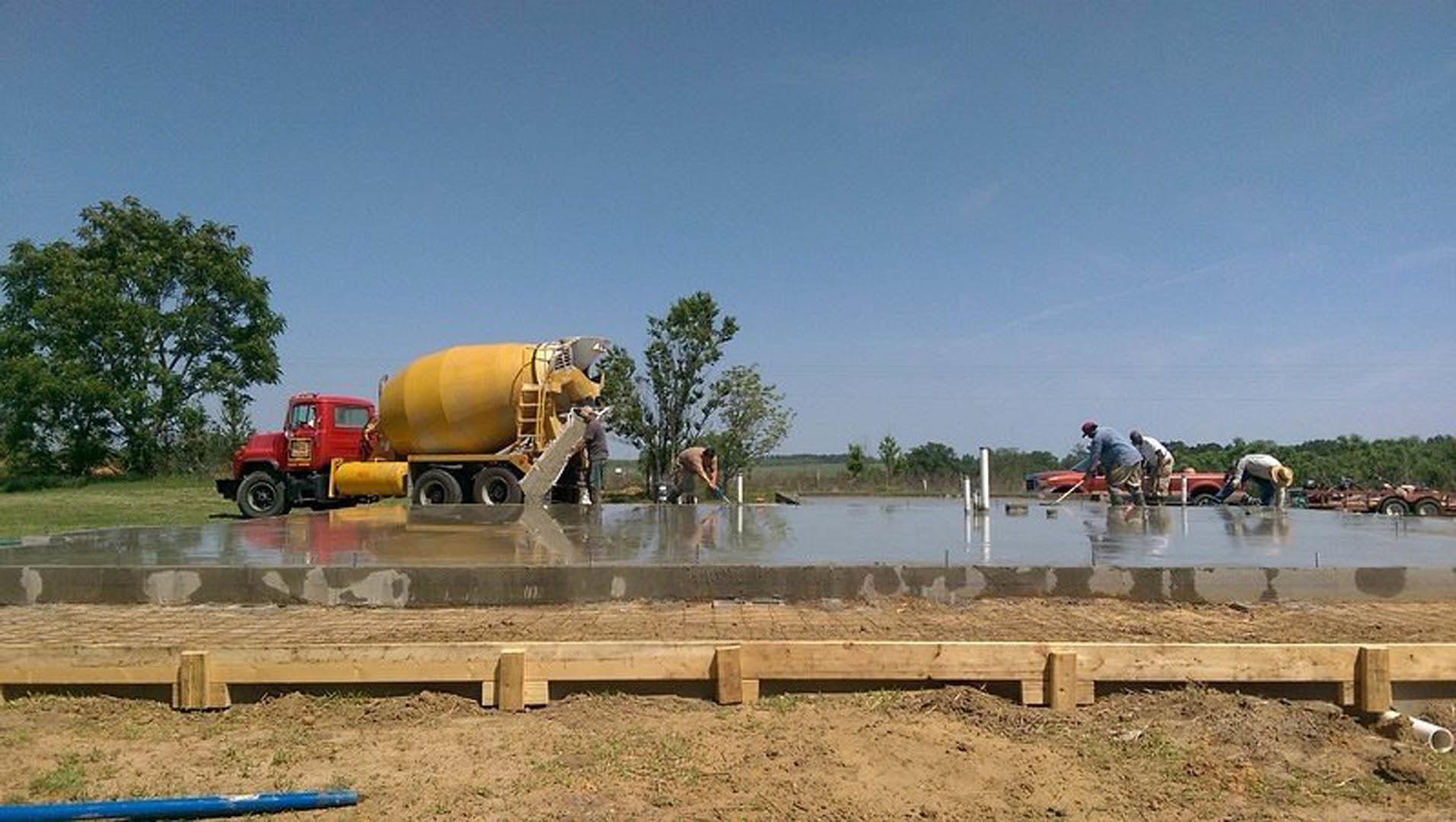 Workers pouring and leveling fresh concrete slab beside yellow truck and concrete mixer, surrounded by grass and trees under open sky