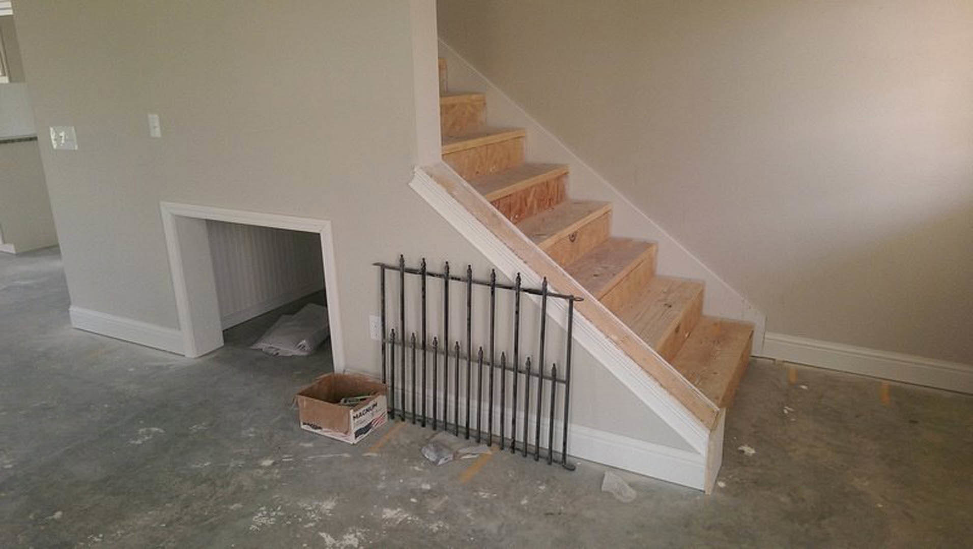 Wooden staircase under construction with exposed steps, metal handrail lying on floor, cardboard box with hole, light switch on plaster wall, white pillow on unfinished flooring