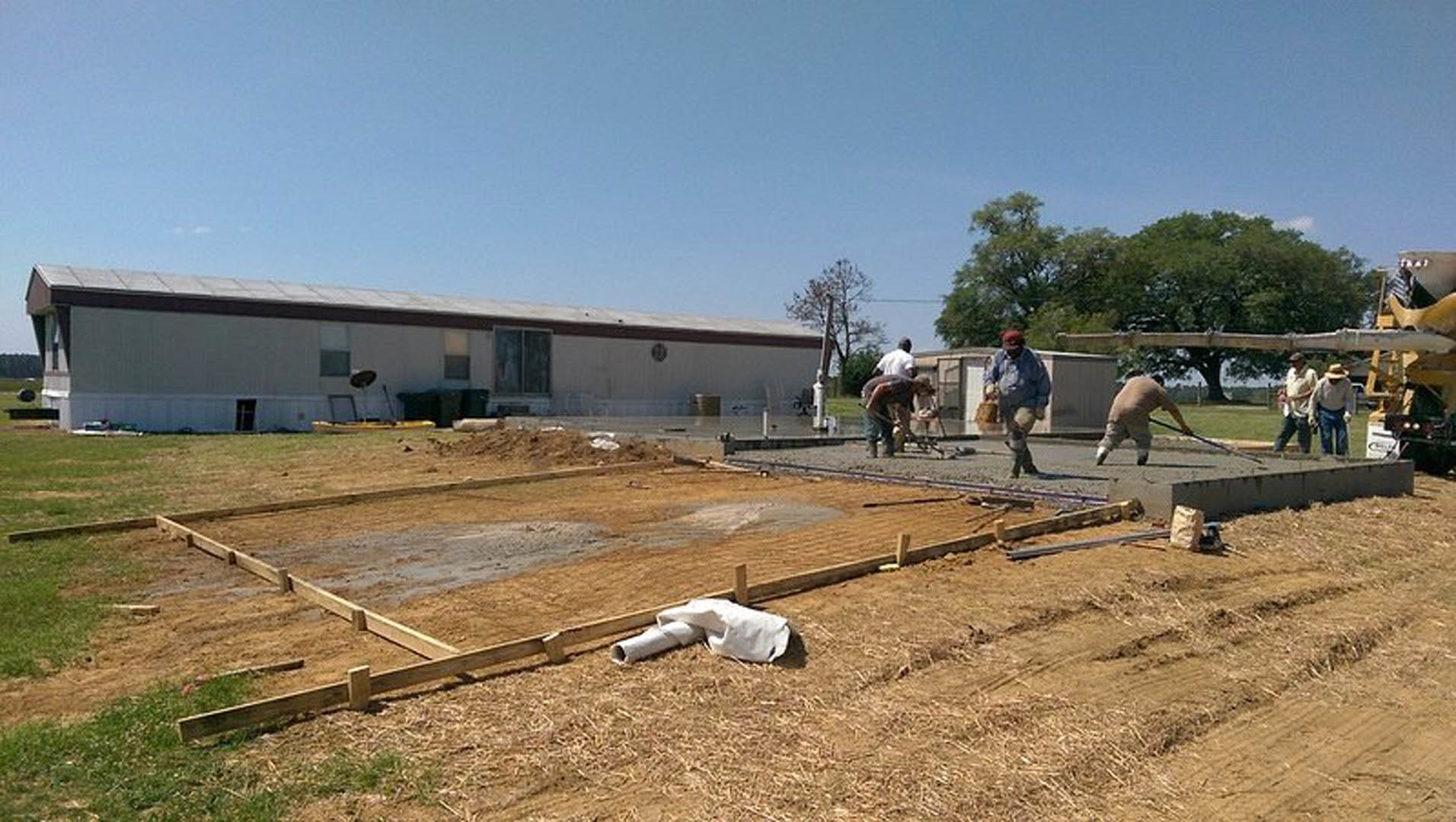 Workers assembling foundation on dirt lot beside partially built house, surrounded by trees and construction materials under open sky