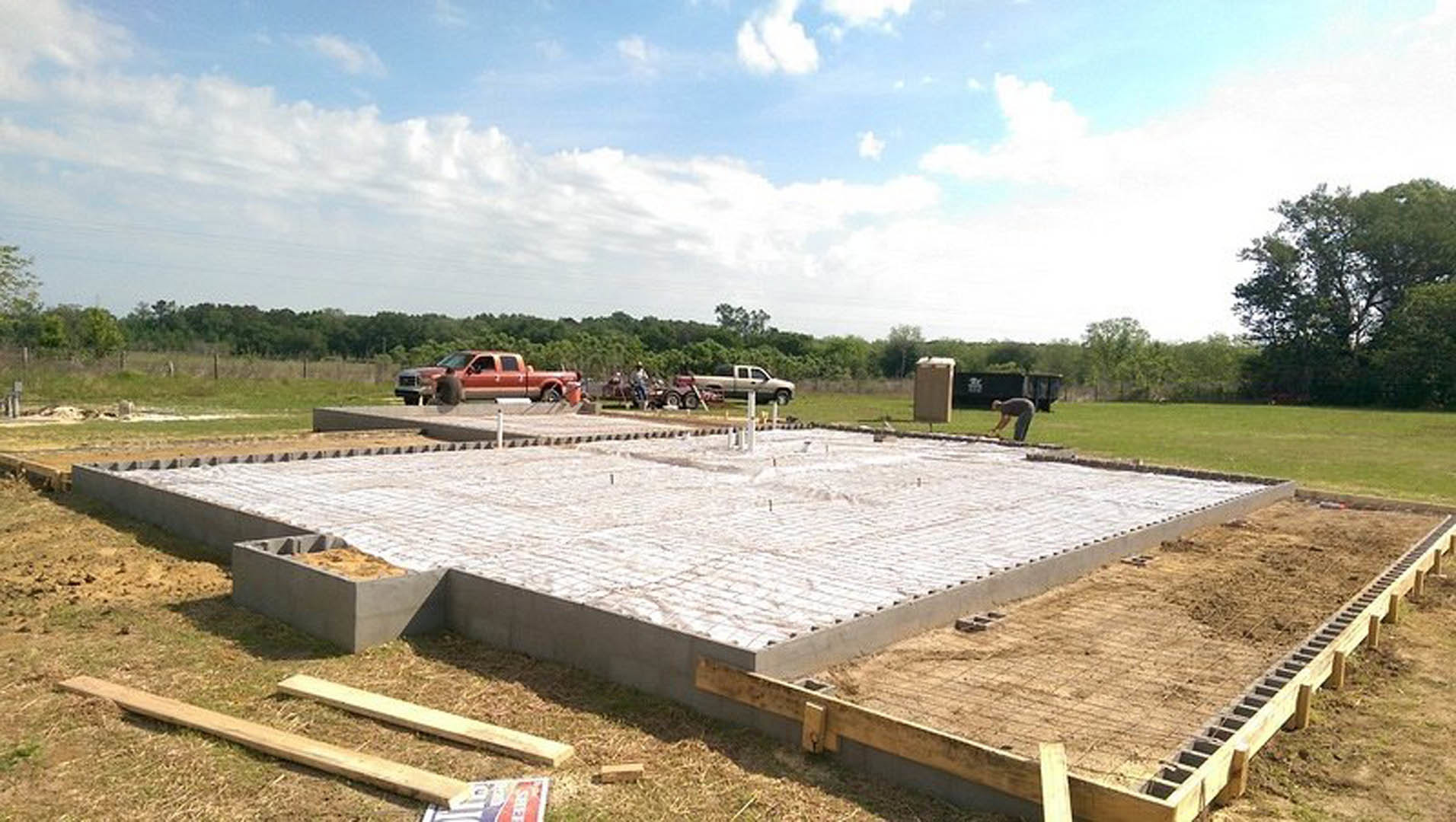 Construction site with two trucks parked on dirt lot, leafy tree in background, blue sky with scattered clouds, grass and exposed foundation visible