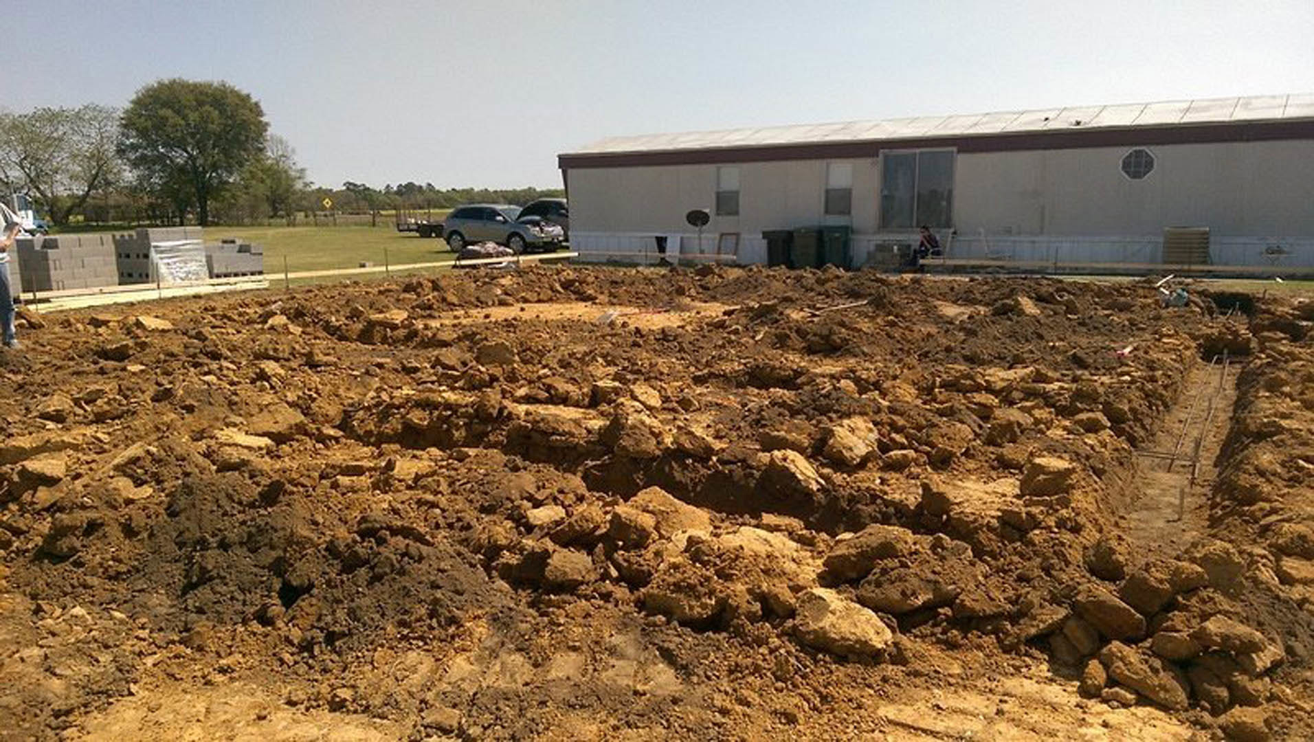 Dirt patch and pile of rocks in front of a house with grid window, leafy tree, person seated outside, and car with open hood on driveway