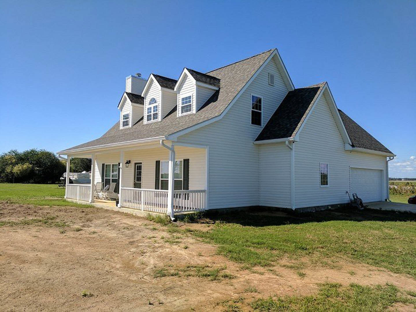 White house with dormer windows and covered porch, dirt path leading to entrance, white fence and trees in background