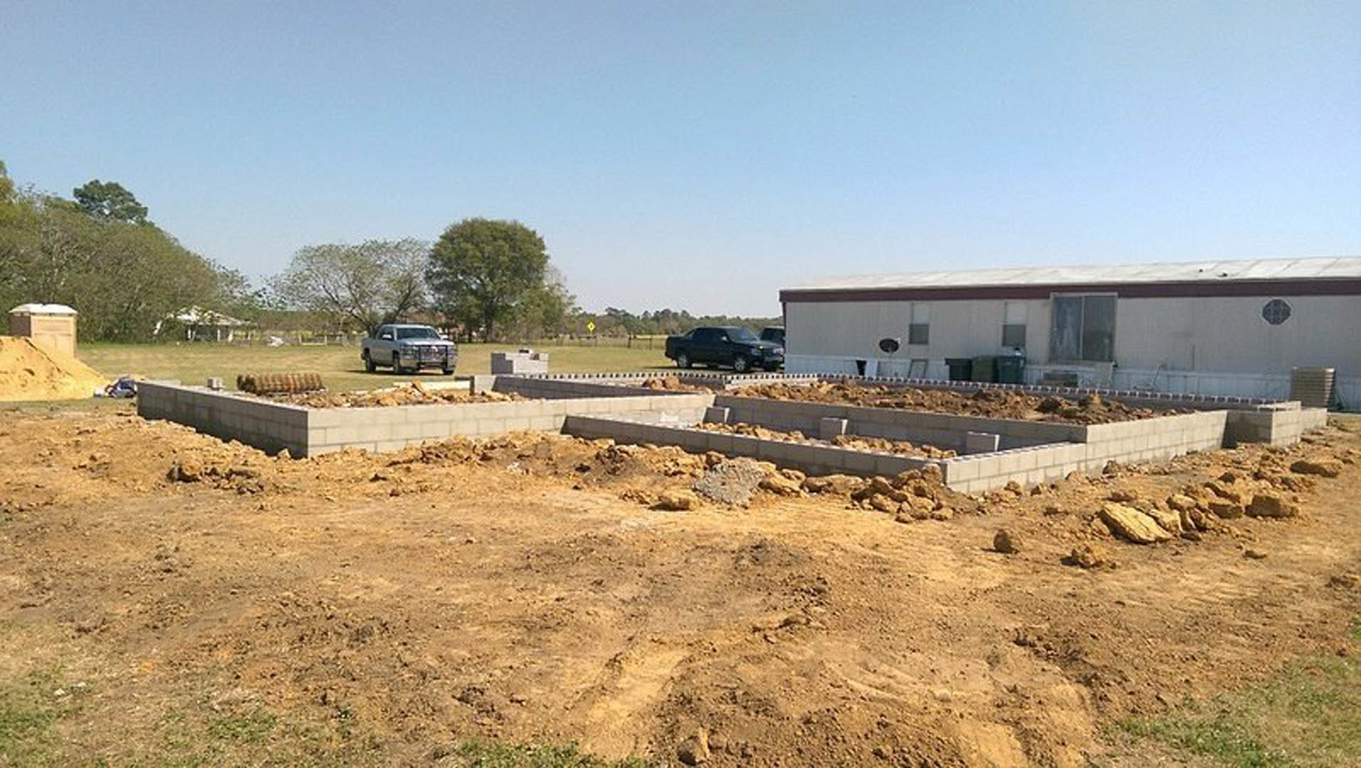 Construction site with exposed soil, scattered concrete blocks, two trucks parked near a white house with open door, trees and grass bordering the lot under a clear sky
