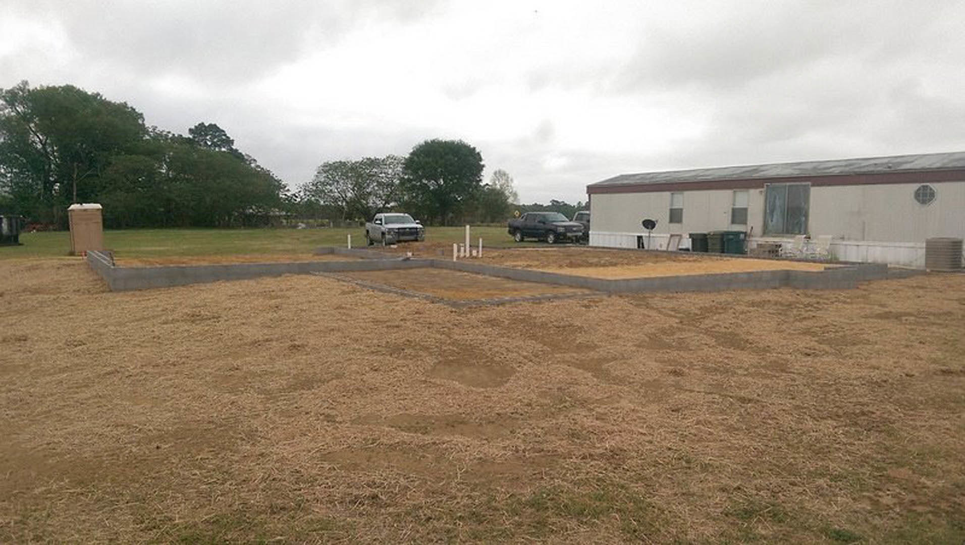 Grey pickup truck parked on gravel driveway in front of white custom home with circular grid window, broken window, and concrete foundation; grassy field and trees in background