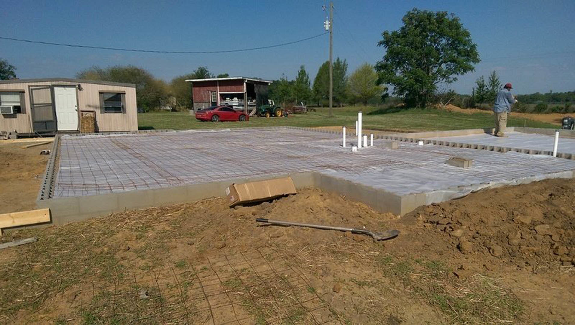 Dirt construction site with a shovel, metal rod, and green-leafed tree beside a grey building with a white door; red car parked nearby and person in blue shirt visible
