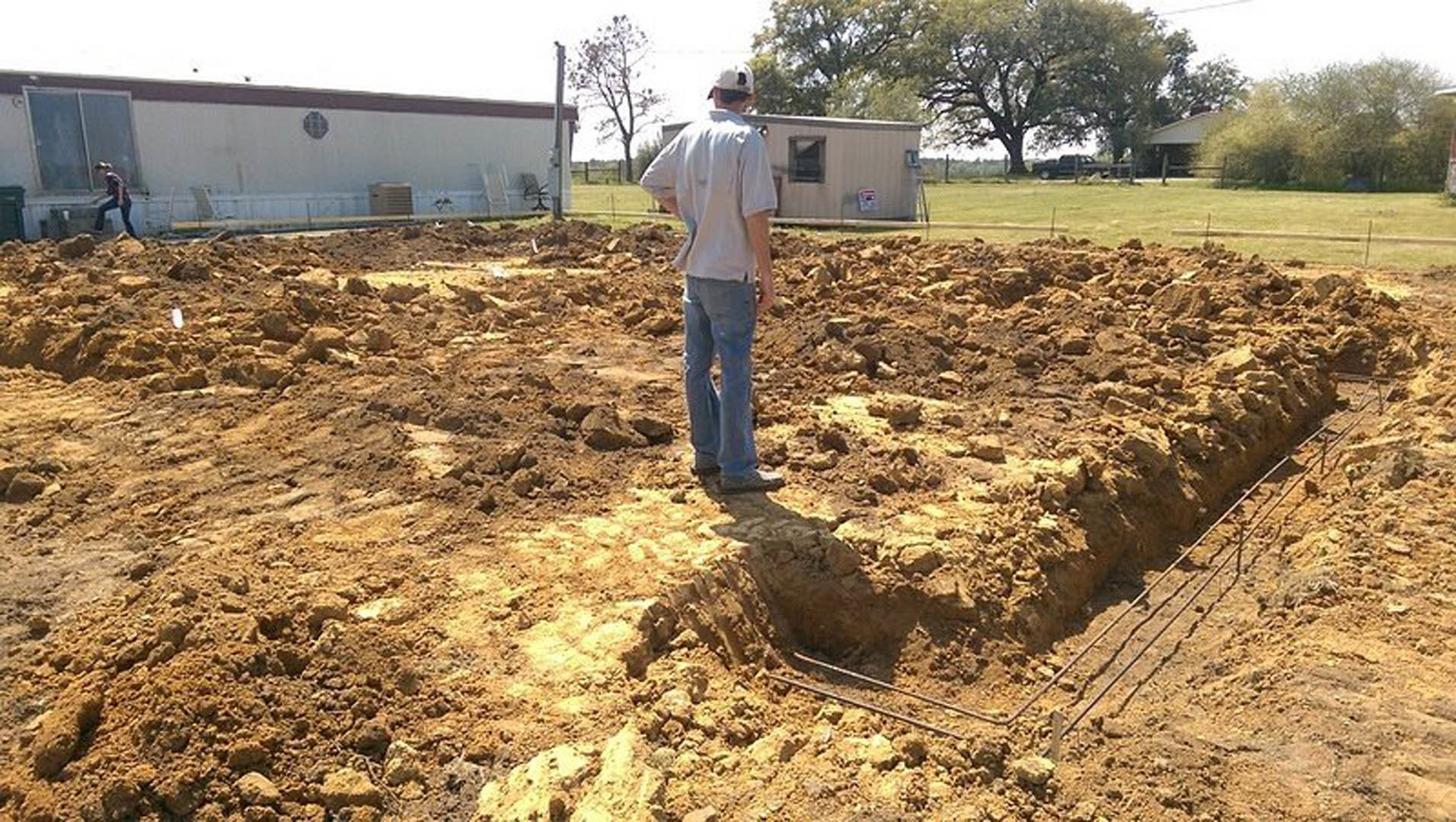 White house exterior with large window, man standing on bare dirt yard, trees and grass in background under blue sky