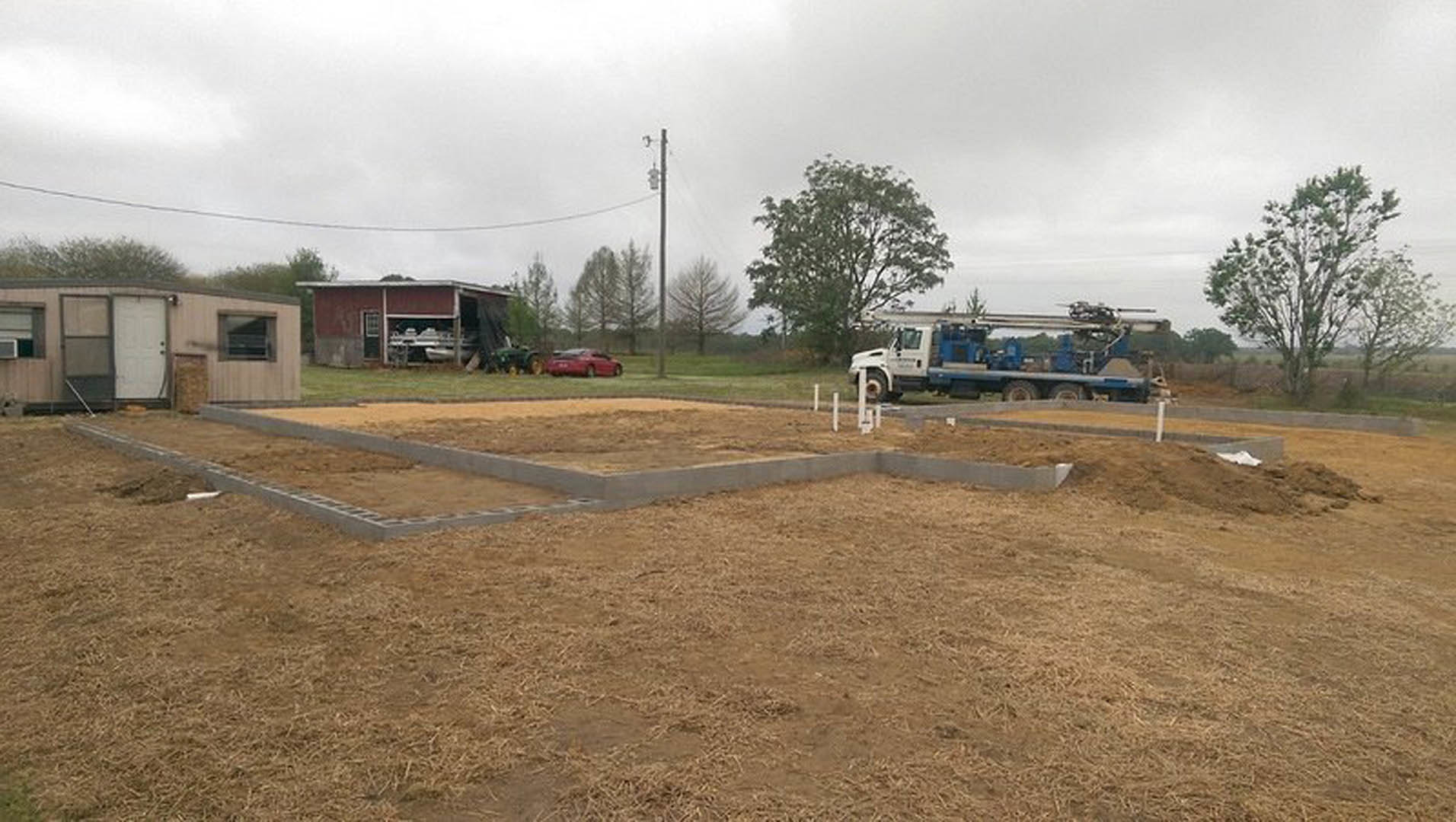 Framed custom home under construction with exposed wood, white door, parked truck on dirt lot, grassy area, utility pole, and scattered building materials