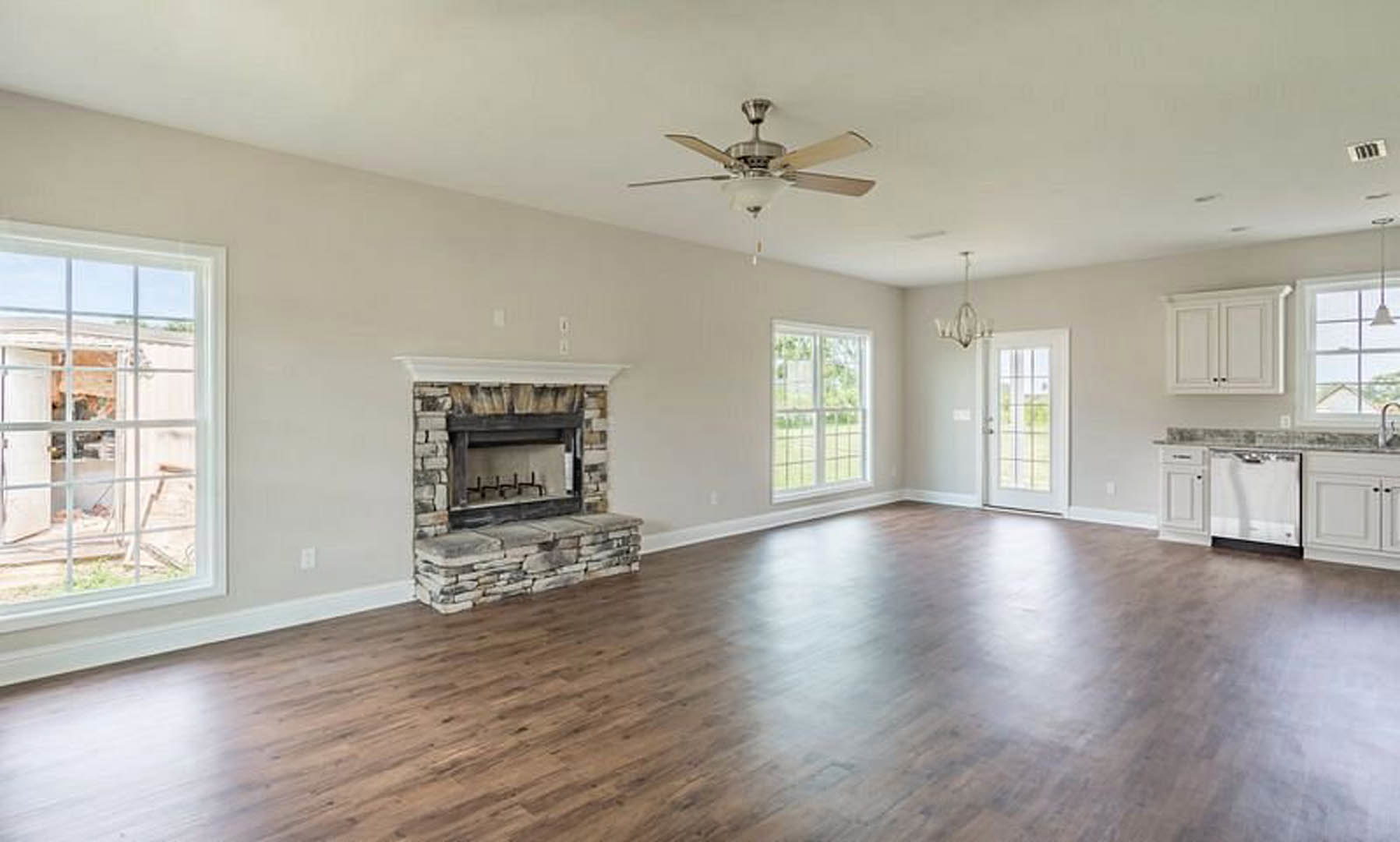 Living room with hardwood floors, stone fireplace framed in wood, ceiling fan with light fixture, large window showing exterior building