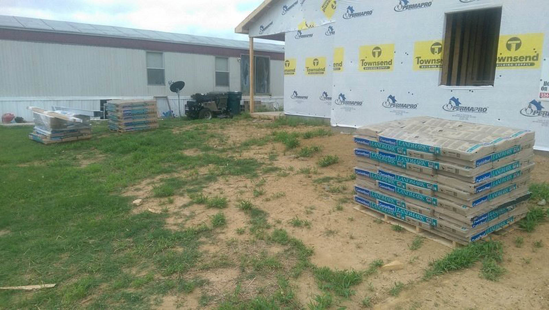 Modern house exterior with white siding, large windows, grassy yard, stacked pallets near entrance, tractor parked in background, satellite dish mounted on wall