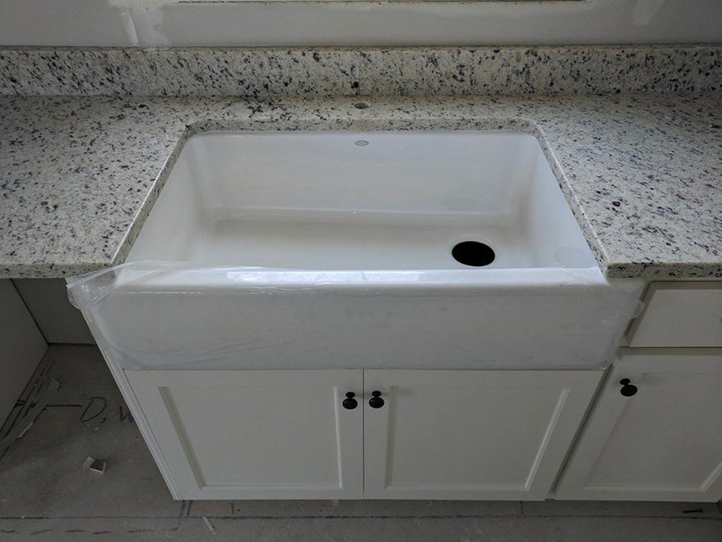 White rectangular kitchen sink with black specks, set into marble countertop, chrome faucet, tile backsplash, and white cabinetry visible.