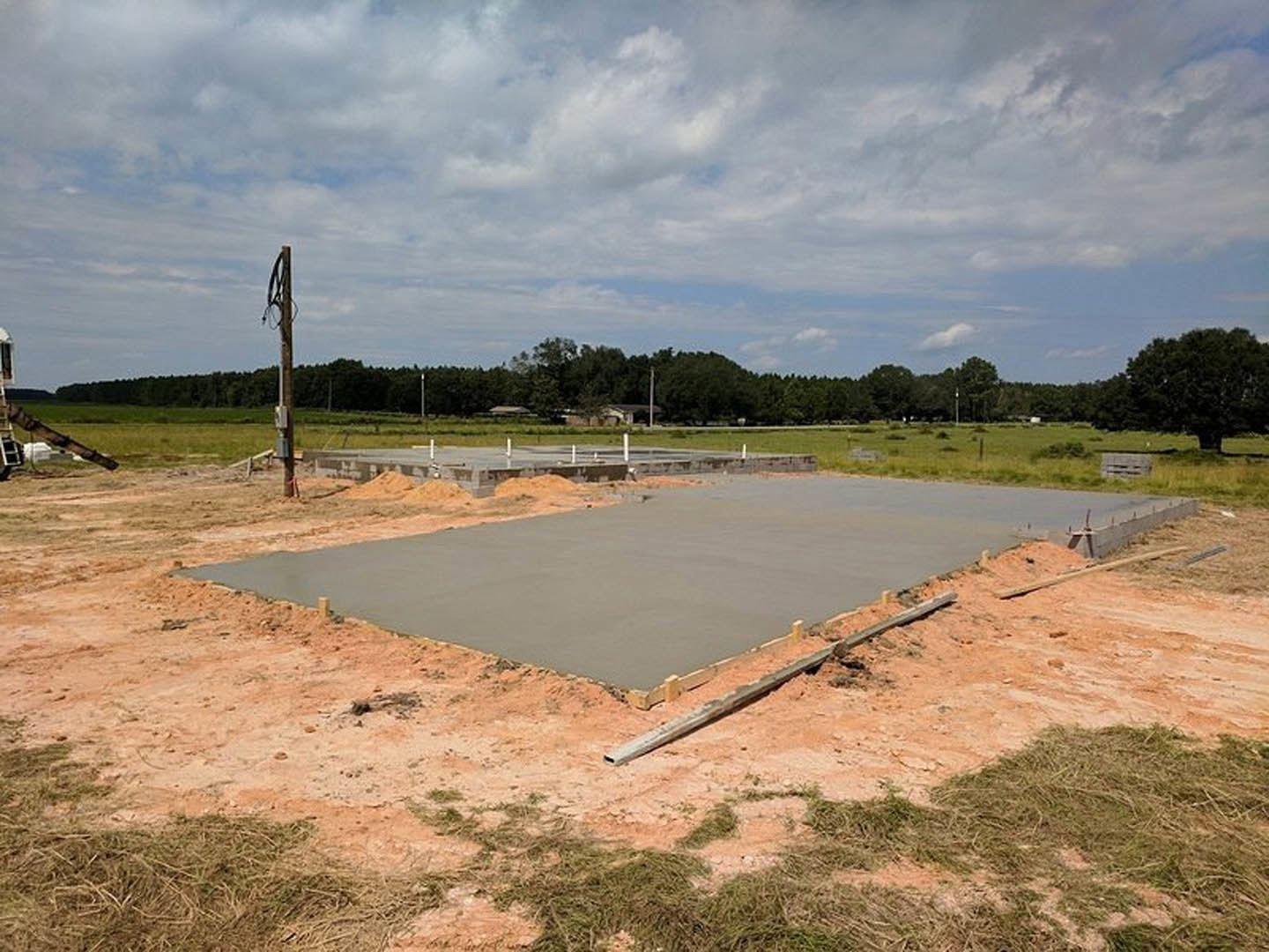 Freshly poured concrete slab foundation on a grassy lot under a cloudy sky, construction equipment and a signpost nearby