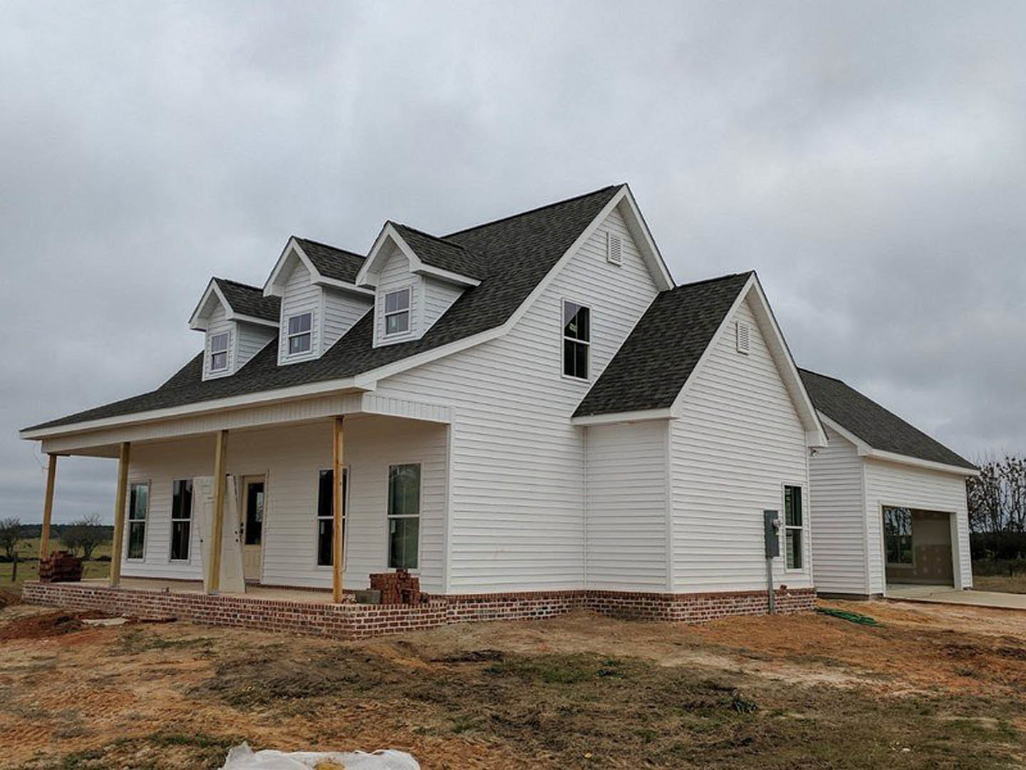 White house under construction with black roof, white-framed windows, brick wall base, patch of grass in front, cloudy sky overhead