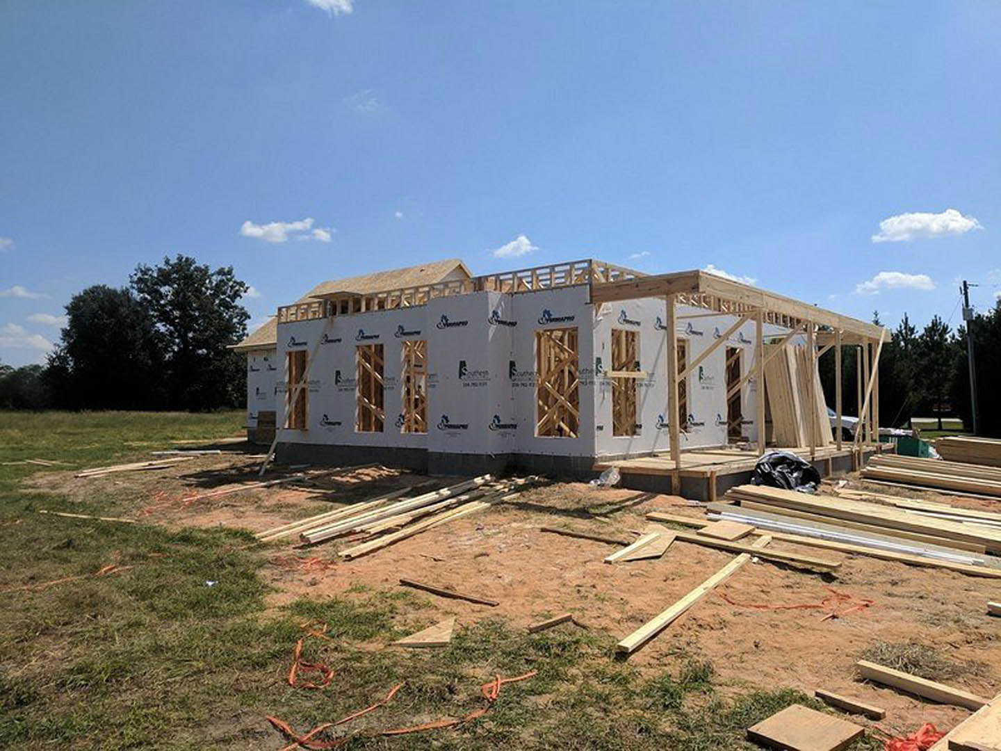 Wood-framed house under construction with exposed beams, plywood sheathing, and trees in the background under a partly cloudy sky